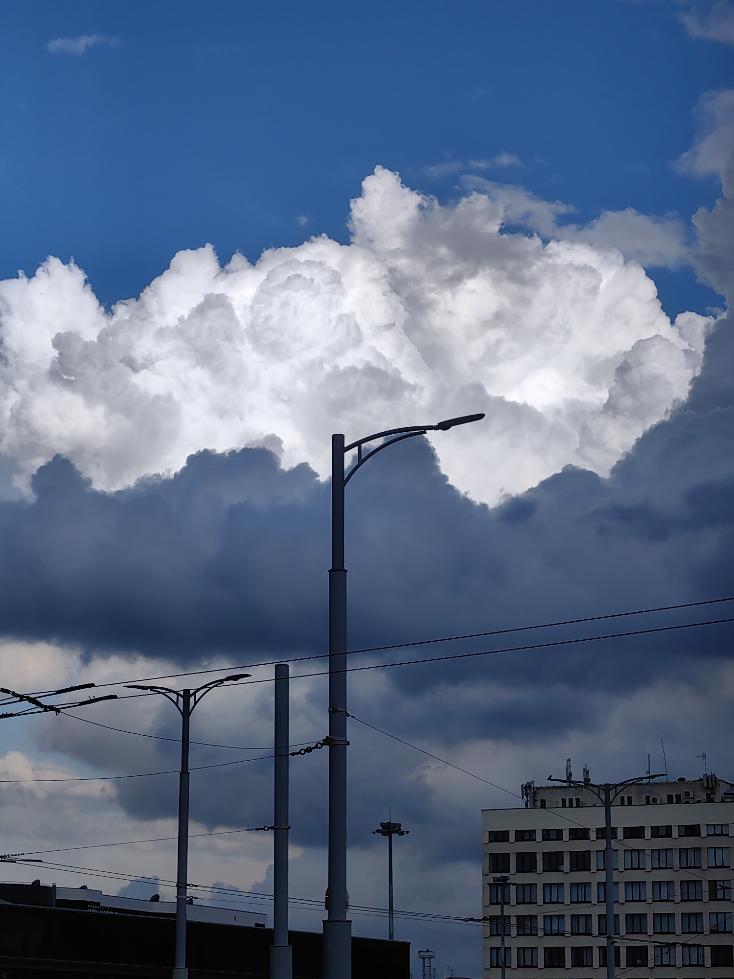 The image captures a dramatic sky over a cityscape in Minsk, Belarus. Large, fluffy white clouds are illuminated by the sun, contrasting sharply with darker, brooding storm clouds below. A modern building with many windows is visible on the right, with streetlights and overhead wires in the foreground, suggesting an urban environment. The scene is likely photographed during daylight hours, possibly before or after a storm.
