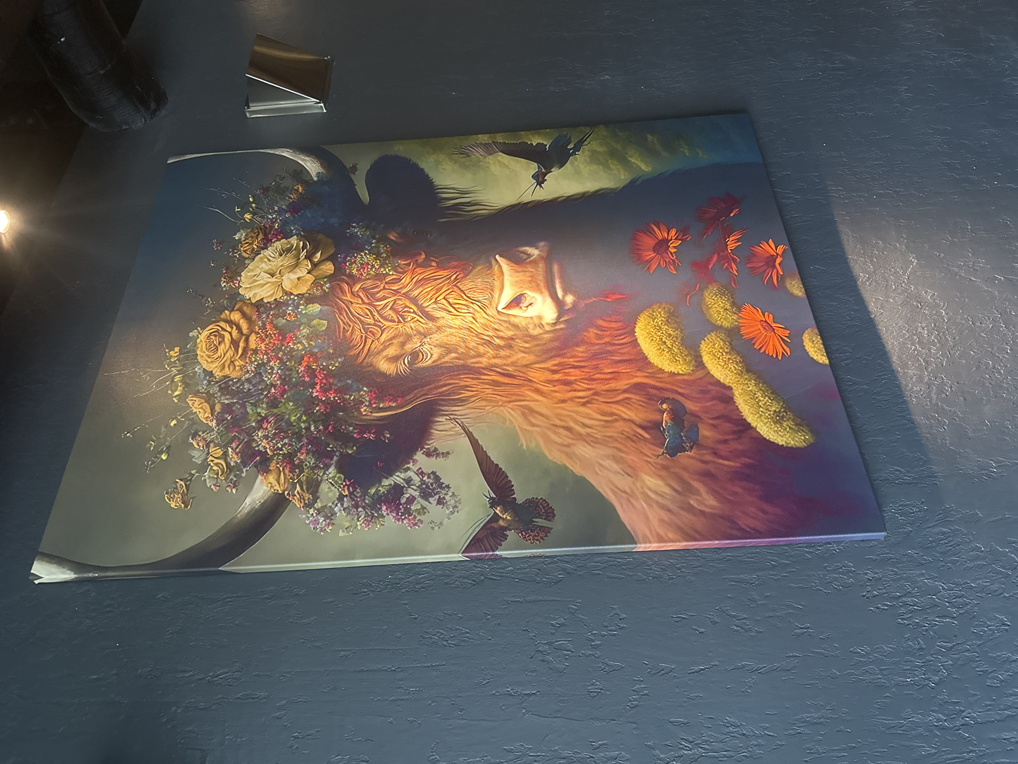 A large, framed canvas art piece depicting a Highland cow adorned with a floral headdress is mounted on a textured, dark blue wall. The artwork features the cow's head and upper body, with its fur rendered in warm, earthy tones and illuminated by directional lighting that casts shadows. A vibrant arrangement of flowers, including roses and smaller blooms in shades of yellow, pink, and purple, crowns the cow's head. Two birds are depicted in flight around the cow. A small, blue butterfly is visible near the bottom left of the canvas. The wall itself has a rough, stucco-like finish. A metallic light fixture is visible on the wall to the right of the artwork. Above and to the right of the canvas, a dark, cylindrical object, possibly ductwork, is visible against the ceiling. The lighting on the wall suggests an indoor setting. There is no information in the image to indicate the location is Moscow, Russia.
