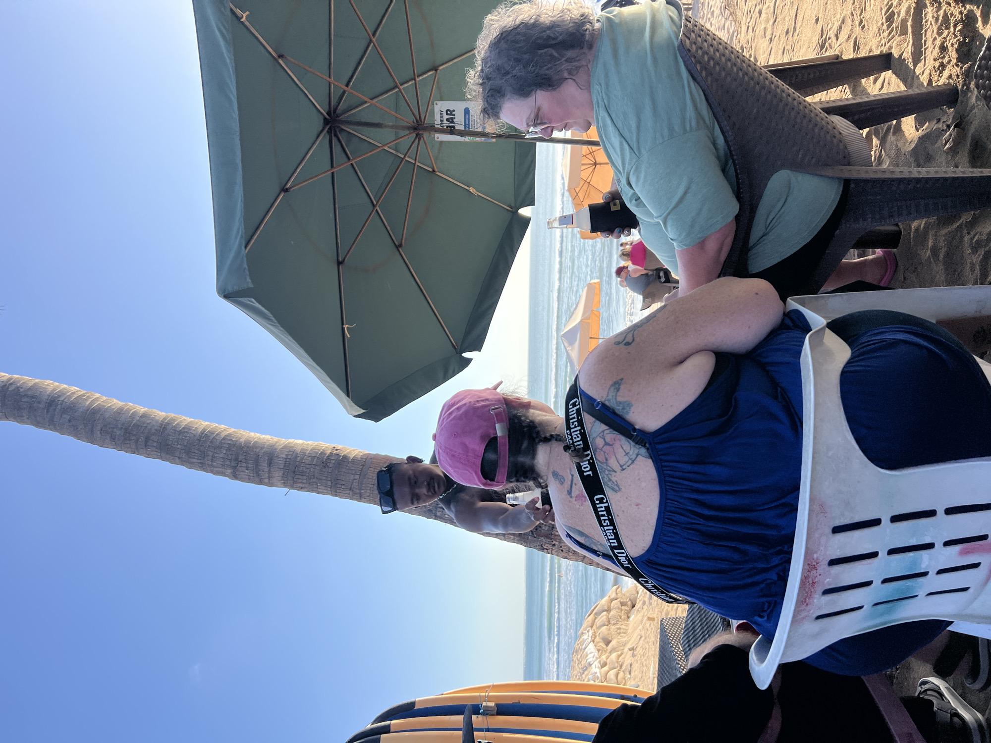 Two women and a man are on a sandy beach in Kuta, Indonesia. One woman is wearing a blue dress and a pink hat, with her back to the camera. She has tattoos on her upper back and shoulders, and a black strap with "Christian Dior" written on it across her chest. The other woman is wearing a green t-shirt and glasses, and is sitting in a wicker chair. She is holding a beer bottle with a black koozie. The man, wearing sunglasses and a black shirt, is standing behind the woman in the blue dress, holding out another beer bottle to her. A palm tree is to the left of the scene, and a green umbrella is overhead. In the background, the ocean and other beachgoers are visible, along with two surfboards.