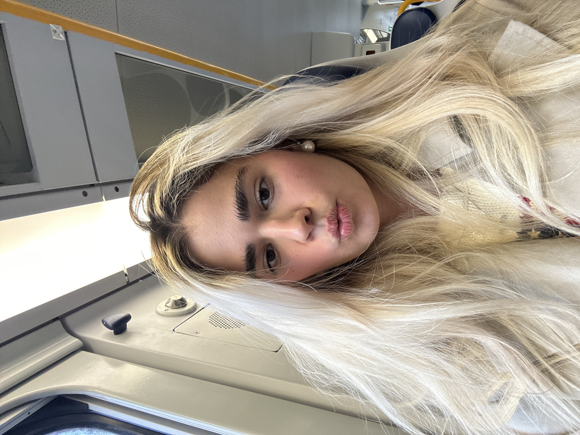 A selfie-style photograph shows a woman with blonde hair, light skin, and dark eyebrows. She is pouting her lips slightly, and is wearing a pearl earring. She appears to be inside a train carriage, based on the interior fixtures such as the window frame with a yellow handrail. The compartment is painted in neutral colors. Due to the presence of the train carriage it is likely that she is in Milan, Italy as she stated.