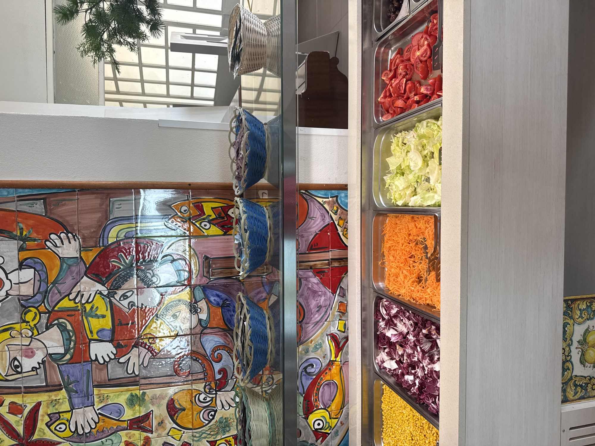 The image shows a buffet with various food items in metal trays. In the foreground, there are trays filled with corn, chopped radicchio, shredded carrots, shredded lettuce, chopped tomatoes, and olives. Above the buffet, there is a shelf displaying three blue woven baskets filled with some unidentified items, and a white woven basket on the right. Behind the shelf, a colorful tiled mural with a folk art style is visible, depicting figures and fish. The buffet is set against a white wall. In the background, a modern window with a grid pattern is seen, and a green plant is placed to the right of the window. The overall setting appears to be an indoor dining area or a restaurant. This buffet setup is consistent with typical offerings at the Hotel Torretta in Capaci, Italy, known for its seaside location and buffet dining options.