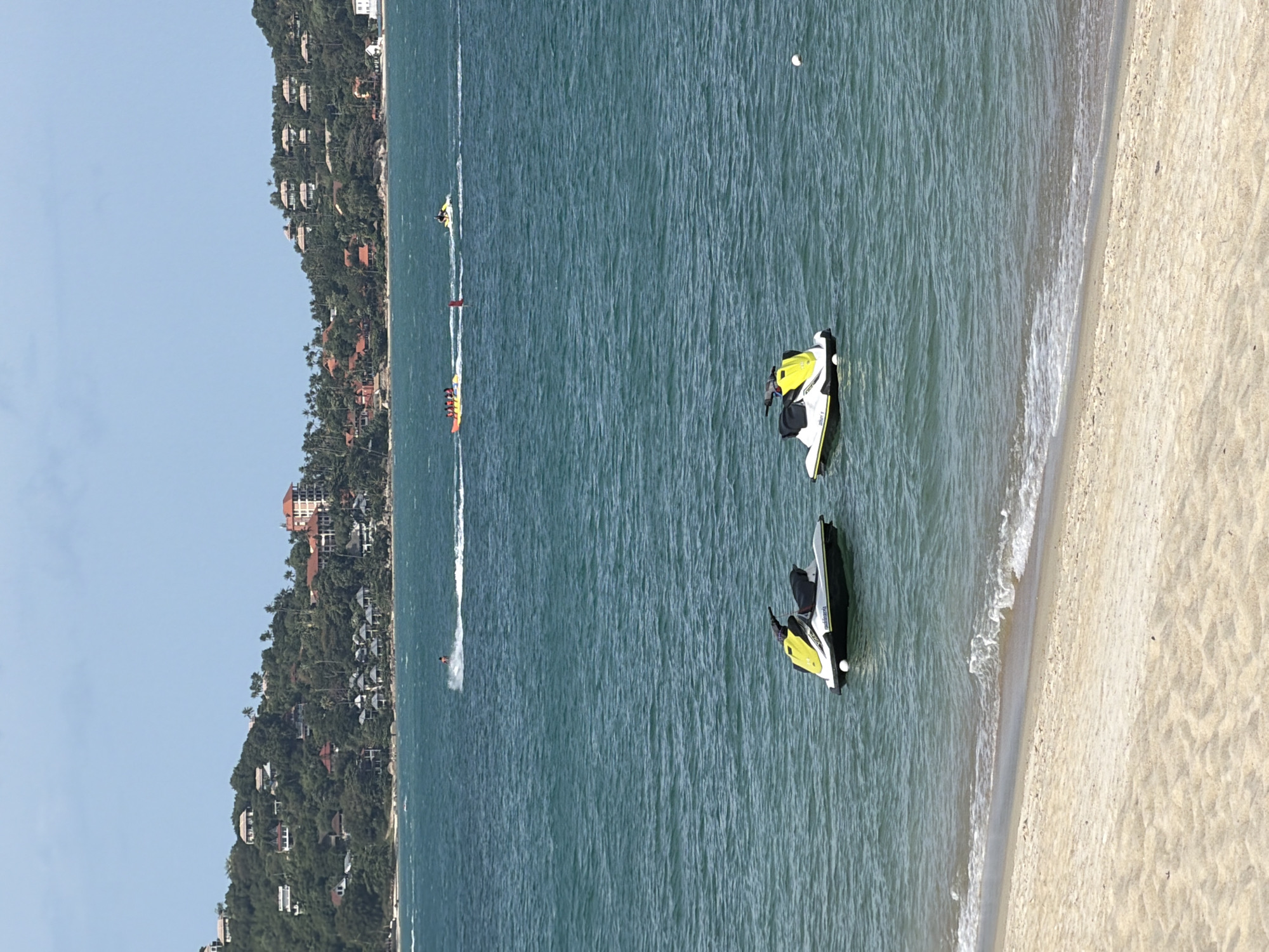Two jet skis are moored in the turquoise water near a sandy beach. The jet skis are white with yellow seats and black handlebars. In the background, a lush green hillside is dotted with houses and palm trees. Several other watercraft, including a banana boat with people and a single jet ski, are visible further out in the bay, along with a person on a jet ski closer to the shore. The sky is clear and blue. This scene is likely located on the coast of Ko Samui District, Thailand.