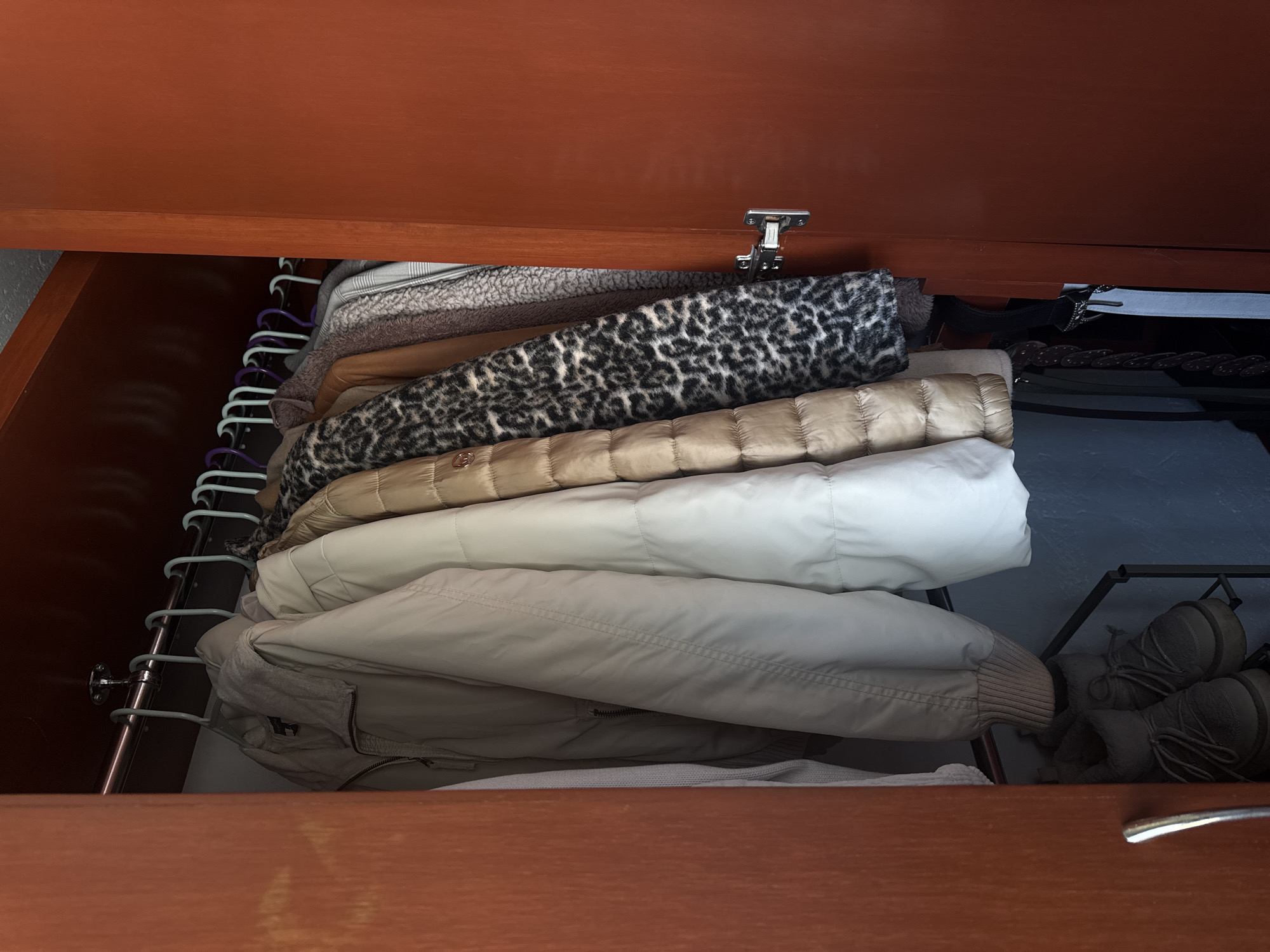 A partially-open drawer reveals a chaotic stack of clothing hanging on a rack. The garments include several jackets, some of which appear to be down-filled and one with a leopard print. Some items appear to be sweaters. The drawer is made of a reddish-brown wood. The setting and location cannot be determined from the image, so the requested location of Heroica Puebla de Zaragoza, Mexico cannot be inferred.