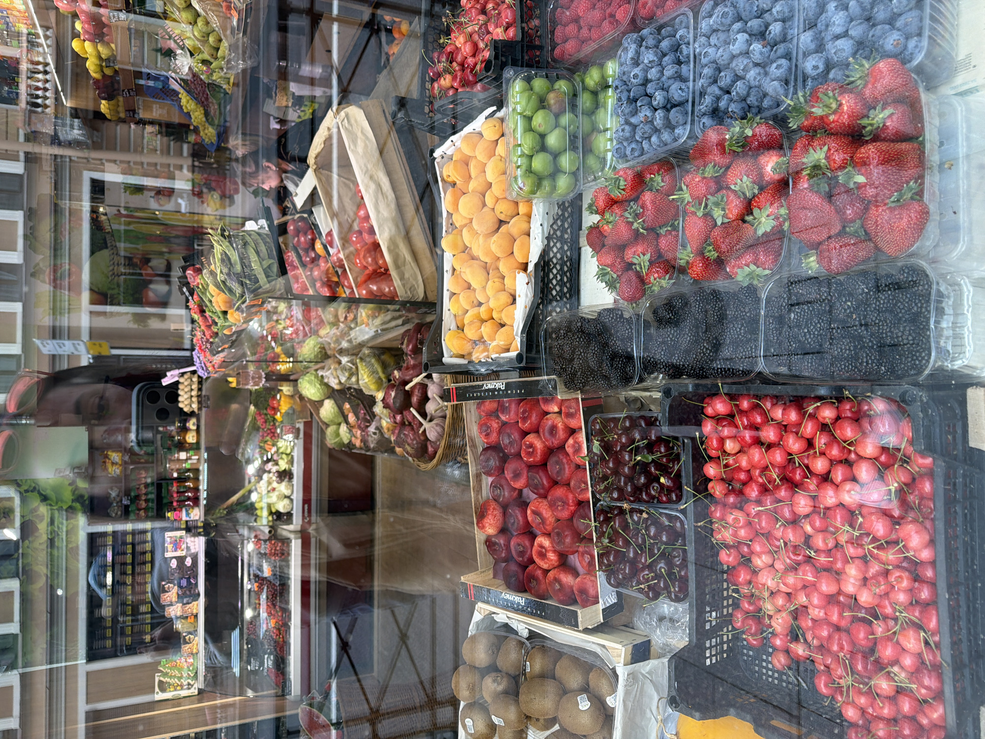 The photograph shows a fruit and vegetable display in a shop window, likely in Saint Petersburg, Russia, based on visible reflections of buildings and signage. The display features a variety of fresh produce, including strawberries, blueberries, blackberries, red and dark cherries, apricots, green plums, red plums, kiwis, tomatoes, garlic, onions, and assorted vegetables. The produce is arranged in plastic containers and crates. The reflections on the glass shop window suggest the presence of buildings across the street, with details such as windows and architectural features visible.
