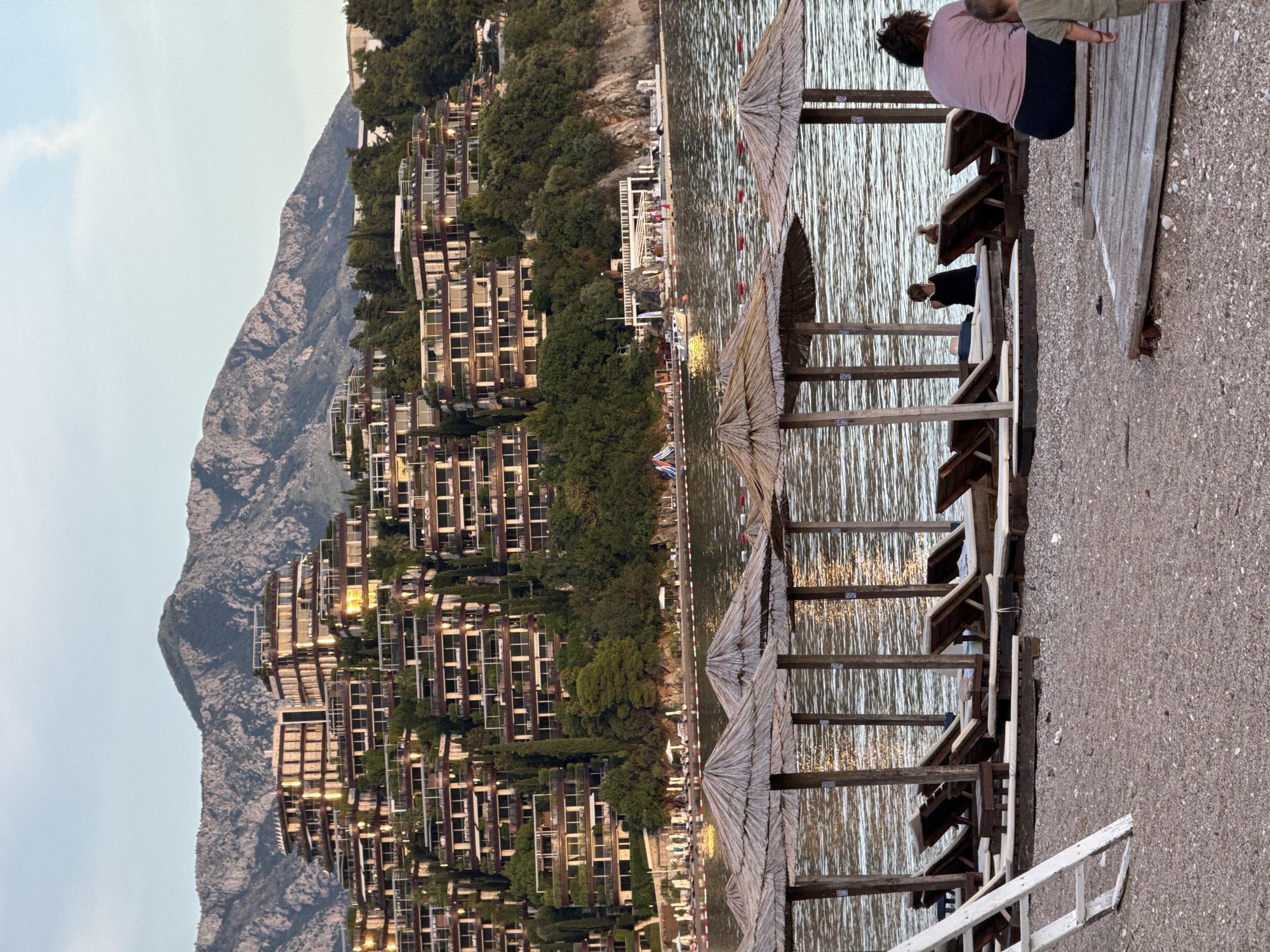 This image depicts a view of the sprawling Aman Sveti Stefan resort complex built into the hillside overlooking the Adriatic Sea in Budva, Montenegro. The resort's terraced architecture, with its numerous balconies and green roofs, is prominently featured against a backdrop of rugged mountains. In the foreground, a pebble beach is set up with straw-covered umbrellas and empty lounge chairs. Several people are visible on the beach and in the water. Two individuals are seated on lounge chairs under an umbrella in the mid-ground, while a woman and a child are seated on wooden decking in the right foreground. The water is calm with gentle ripples reflecting the light. The overall scene captures a serene evening at a coastal resort.
