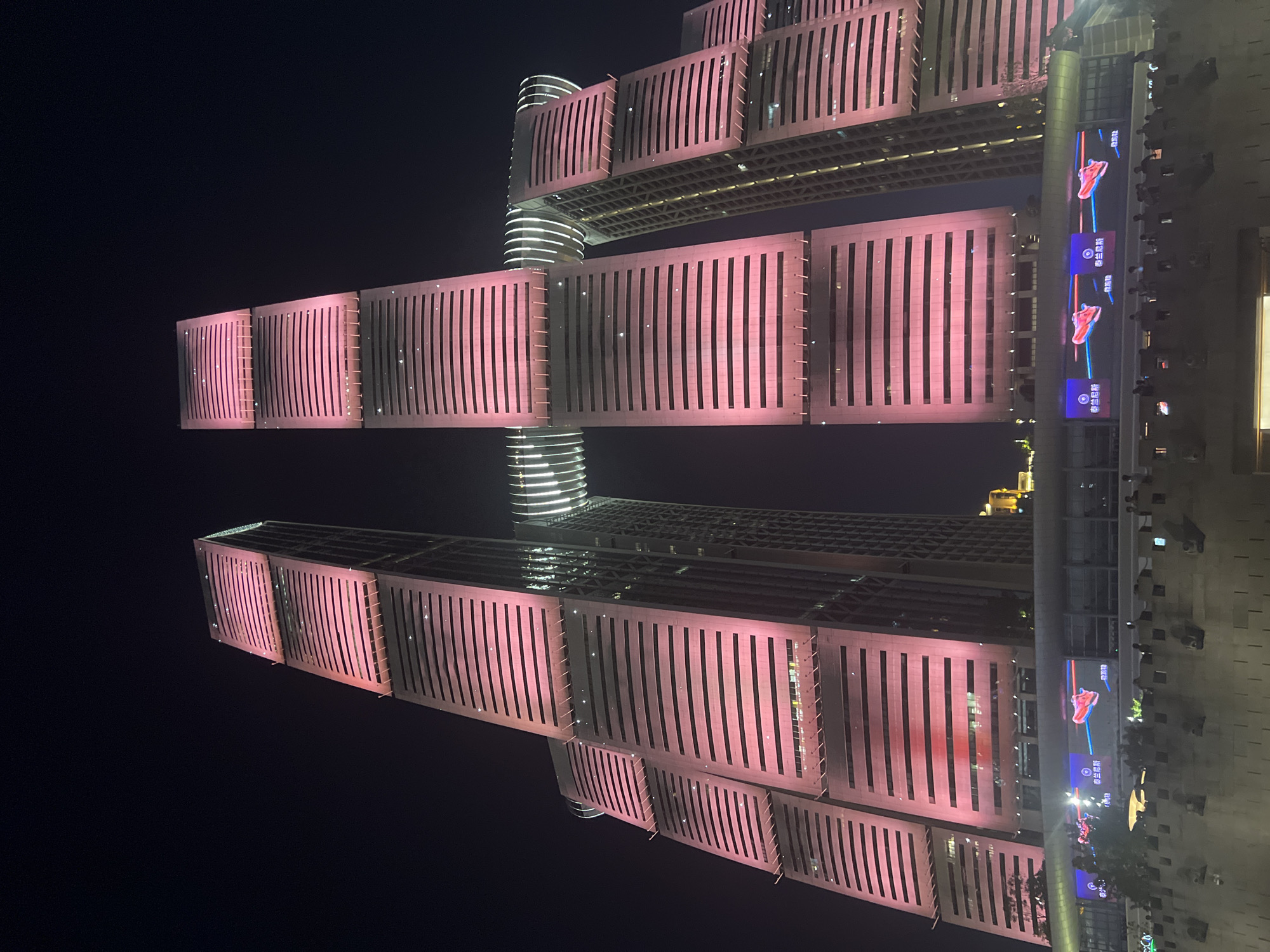 This is a low-angle, nighttime photograph of the iconic Twin Towers of Nan'an District, China, also known as the "Belt and Road Tower." The towers are illuminated with a vibrant pink light, highlighting their distinctive horizontal window bands and stepped architectural design. The towers are connected by a curved, illuminated skybridge. In the foreground, a lower building with a large LED screen displays advertisements featuring images of red athletic shoes. Silhouetted figures of people are visible on a walkway in front of this lower building, looking towards the towers or the screen. The overall environment is urban and modern, with the dark night sky providing a stark contrast to the illuminated architecture.