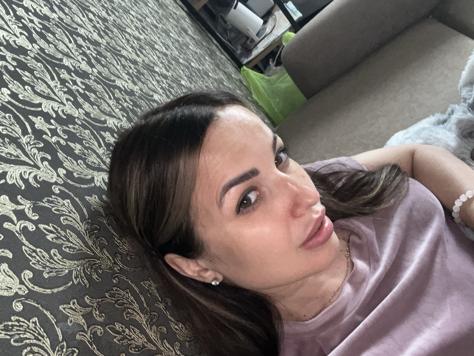 A woman with long, dark hair and brown eyes is shown in a close-up, overhead shot, looking at the camera. She is wearing a light purple t-shirt and a delicate necklace. A sparkly earring is visible in her left ear. Her skin appears smooth and her lips are slightly parted. In the background, a patterned wallpaper with floral motifs is visible. A portion of a sofa and some objects on a shelf, including a green plastic bag, are also in the frame. A white, fluffy object, possibly a small dog, is partially visible in the lower left corner. The scene appears to be indoors, in an unknown location.