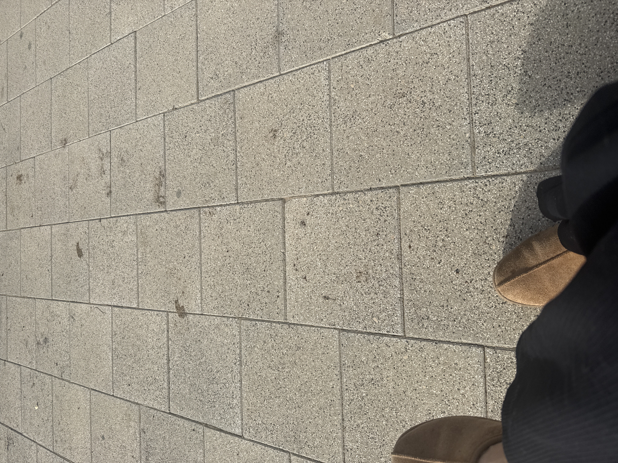 A close-up, angled shot shows a paved sidewalk in Tel Aviv, Israel, composed of light gray rectangular stones. A person is standing on the sidewalk, wearing brown slippers and dark-colored clothing covering most of the body except for the feet and the edges of the slippers. The sidewalk stones are laid in a brick-like pattern, and some have minor stains or discolorations. The lighting suggests daylight.