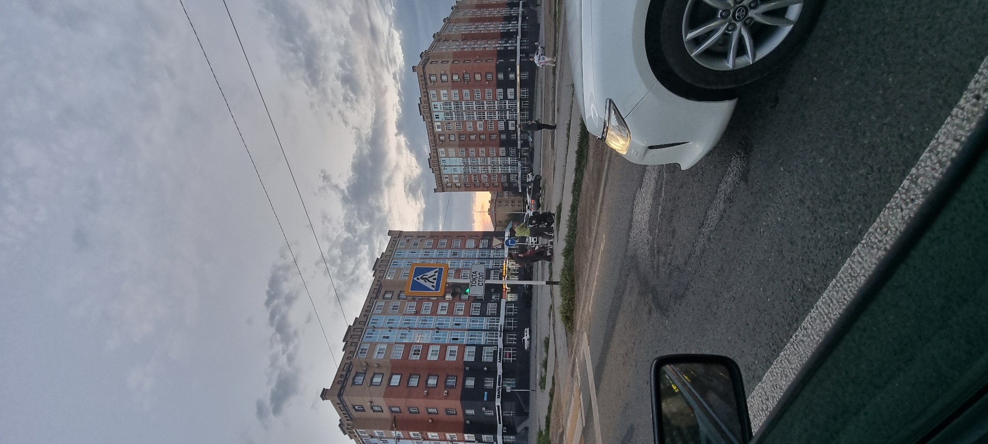 The image captures a street scene in Aktobe, Kazakhstan, likely during an overcast evening, with a sunset visible in the background. Two multi-story residential buildings, characterized by brick facades with sections of blue-tinted windows and dark lower levels, dominate the skyline. In the foreground, a white car is partially visible on the right side, with its front bumper, headlight, and wheel in focus. The street is paved asphalt with visible lane markings and a crosswalk. A traffic light with a green signal is illuminated. A pedestrian crossing sign is mounted on a pole. Several individuals are present near the crosswalk, some pushing strollers, suggesting a residential area. The sky is filled with dramatic clouds, typical of a stormy or sunset sky. The overall impression is a typical urban environment in Kazakhstan.