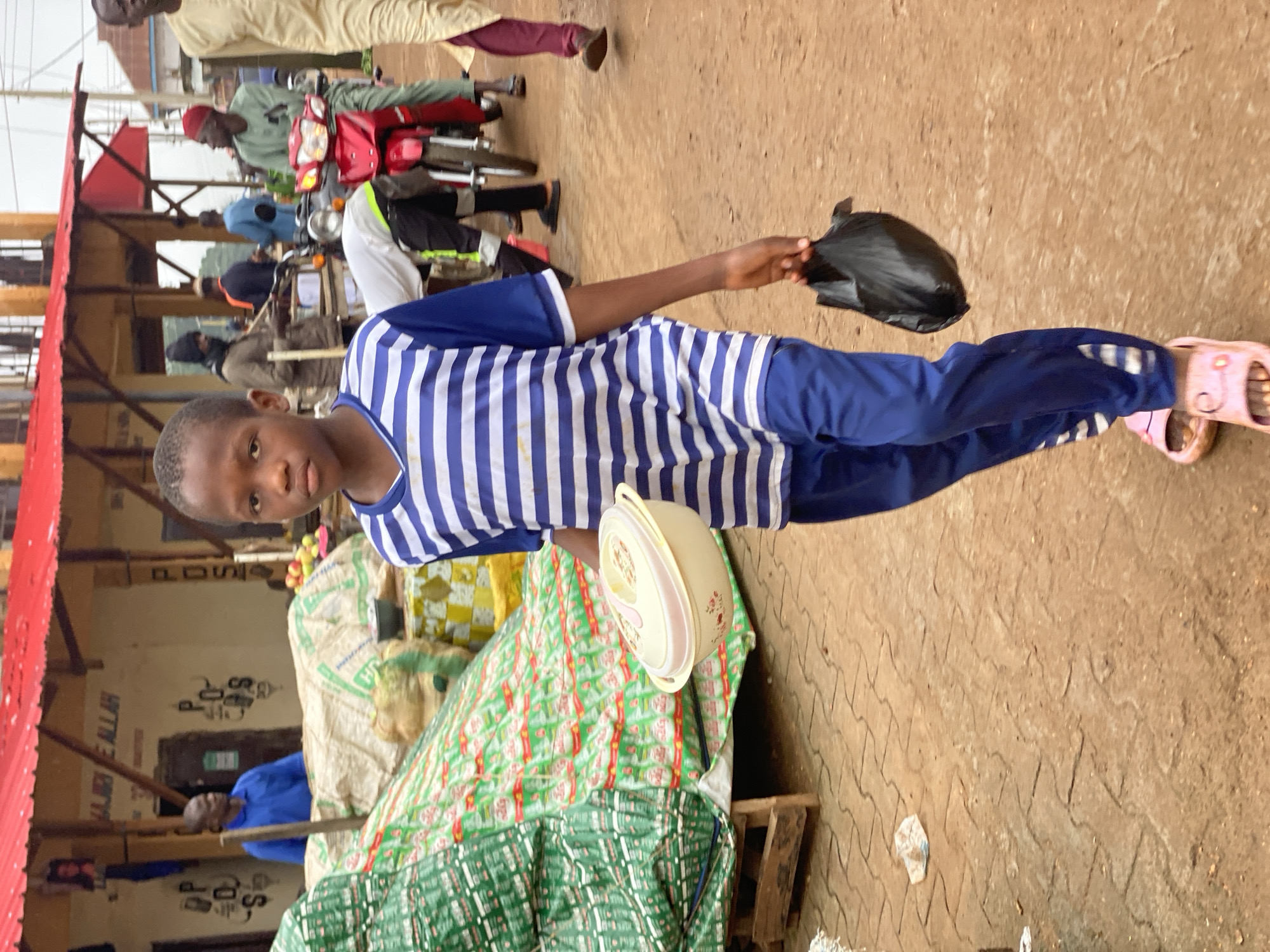 In Kafanchan B, Nigeria, a young boy walks through a marketplace, holding a plastic food container and a black plastic bag. He is wearing a blue and white striped t-shirt and blue pants, with pink sandals on his feet. In the background, a red motorcycle is visible, and several other people are present in the market. Stalls with goods are set up, covered by green and white tarpaulins. The ground is made of reddish-brown interlocking paving stones.