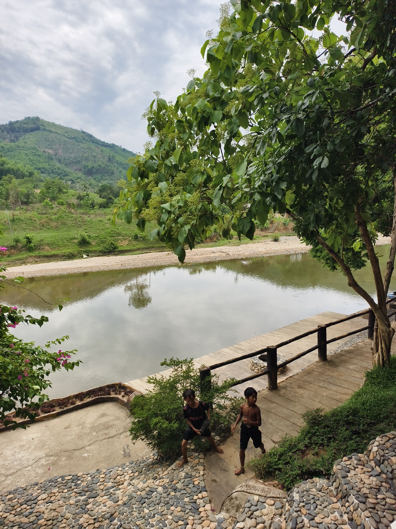 Two young boys stand on a concrete and cobblestone patio adjacent to a wooden deck with a rustic railing. The patio overlooks a calm river reflecting the cloudy sky. Lush greenery frames the scene, with a prominent tree on the right and smaller bushes scattered around. In the background, rolling hills and mountains are visible under the cloudy sky. Based on the information provided, the location is Nam Khanh Vinh, Vietnam.