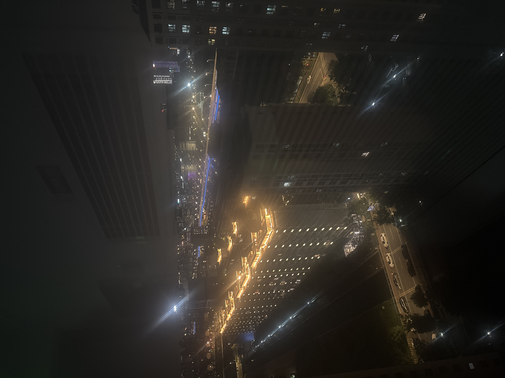 The image depicts a nighttime aerial view of a city, likely in Suzhou Industrial Park, China, characterized by an urban landscape illuminated by artificial lights. In the foreground, a long, multi-story residential building dominates the left side, with numerous windows lit from within. Across a street from this building, another large, modern building stretches diagonally across the frame, its upper floors featuring bright, illuminated decorative elements.
Further into the background, a complex network of elevated roads and bridges, possibly a highway interchange, is visible, marked by lines of streetlights. Distant skyscrapers and other urban structures are scattered throughout the scene, their lights creating a hazy glow in the night sky. Several cars are present on the roads, their headlights and taillights faintly visible. The overall impression is of a bustling, modern city at night, with a sense of depth and scale conveyed by the arrangement of buildings and infrastructure.