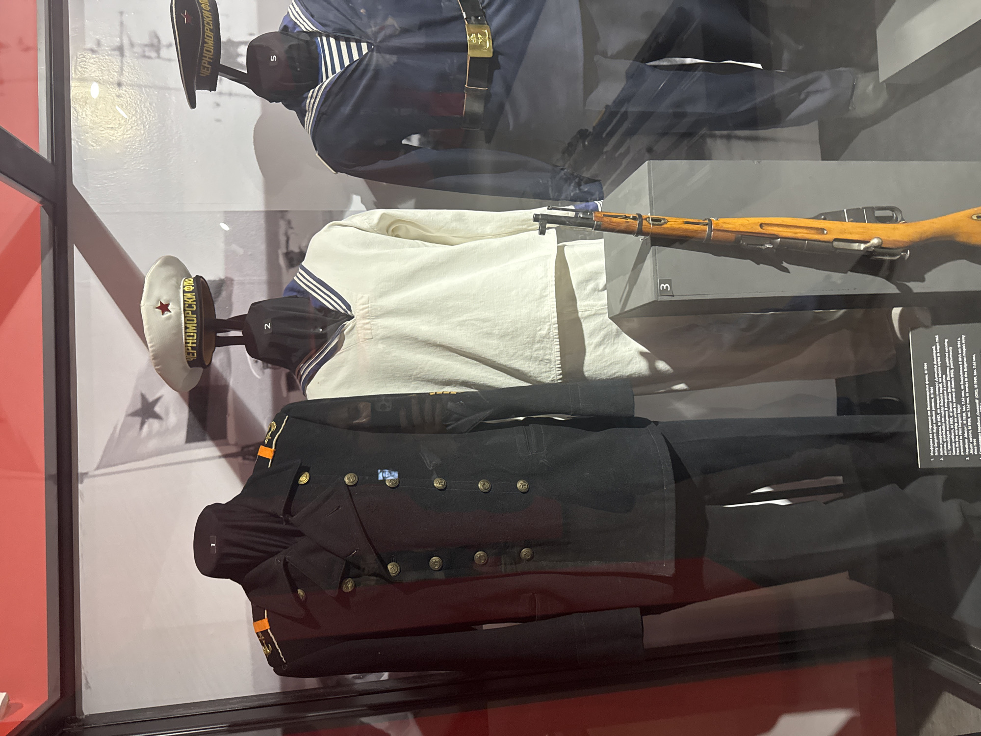The image shows a display case in what is likely a museum. In the case are several types of uniforms, most likely of Russian origin. A hat with a red star and the words "ЧЕРНОМОРСКИ ФЛ" (CHERNO MORE SKI FL), translating to Black Sea Fleet, rests atop a mannequin head. Beneath it is a white and blue striped shirt. Below this is a dark navy blue jacket and pants with golden buttons, and what appears to be a ribbon. Above the hat is another navy uniform, and between the two is a rifle, also resting on a display. A small plaque sits next to the rifle. The background of the case appears to be a blown-up picture of a navy ship or base, with a large red star. The overall scene suggests a historical or military exhibit in Sofia, Bulgaria, showcasing uniforms and equipment, specifically from the Black Sea Fleet.