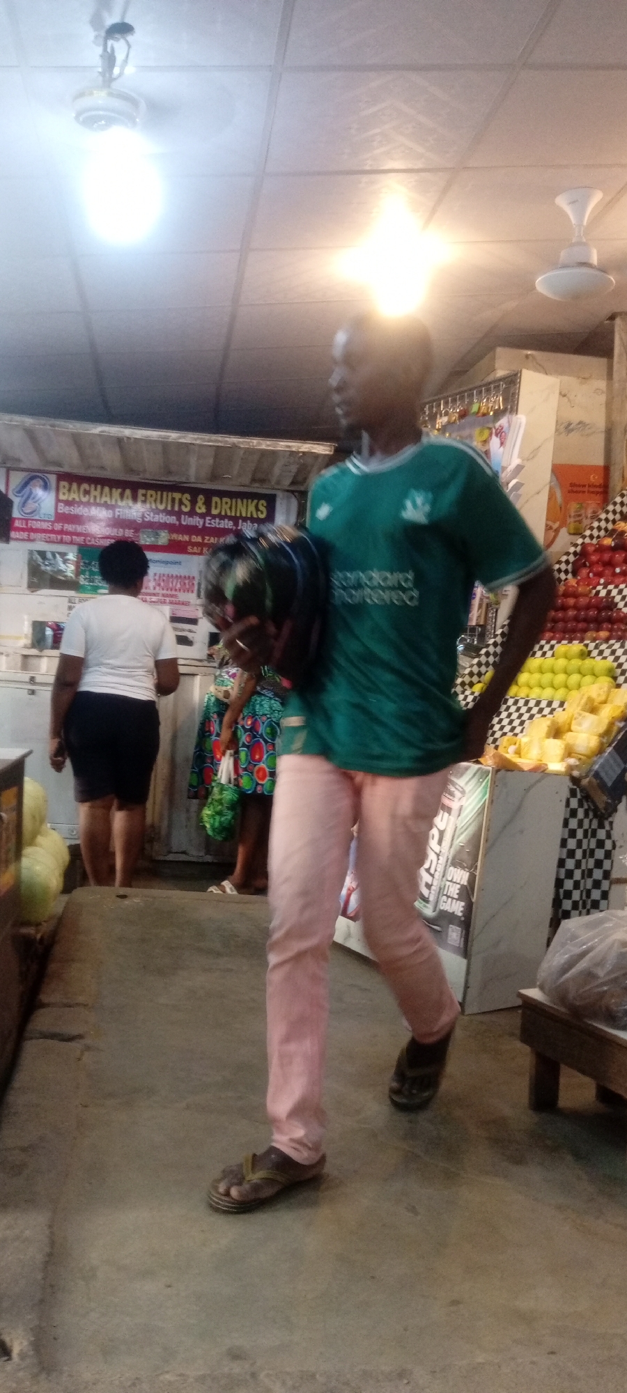 The image captures a candid moment inside a fruit and drinks store in Kano, Nigeria. The store is identified as "Bachaka Fruits & Drinks," located beside the Miko Filling Station in Unity Estate, Jaba. A man wearing a green "Standard Chartered" Liverpool football club jersey and pink pants walks towards the foreground, holding a dark, possibly watermelon-like object. He is wearing flip-flops. Other customers are present, including a woman in a white top and black shorts standing near a refrigerated display case filled with produce. Fruits such as oranges and apples are stacked on shelves in the background. An advertisement for "Own the Game" is visible. The lighting in the store is bright, with two ceiling fans providing ventilation. The floor is made of concrete.