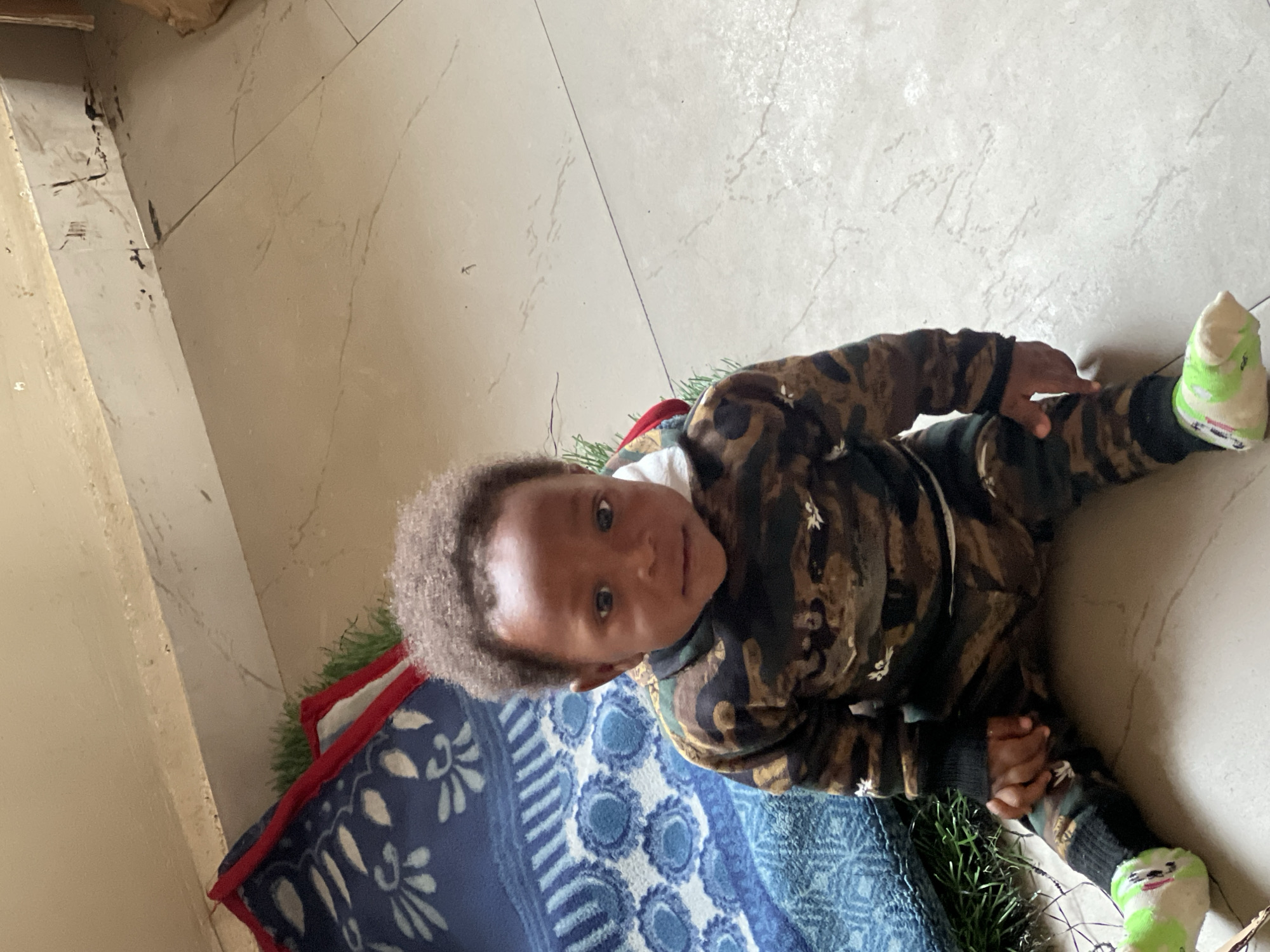 A young child is depicted in an indoor setting, lying or sitting on a tiled floor in Jos, Nigeria. The child, appearing to be a toddler, has dark skin and short, curly dark hair, and looks directly at the camera with a calm, curious, and slightly smiling expression. They are dressed in a camouflage-patterned long-sleeved top and matching pants, suggesting a warm tracksuit. Light green and white patterned socks cover their feet.
The child's body extends partially onto a blue blanket or mat, which features white, possibly floral or geometric, patterns and a red trim. Next to the blanket, a patch of green, spikey material, resembling artificial grass or a festive garland, is visible. The floor consists of large, light-colored tiles with subtle dark veining. In the top left corner, a light-colored wall or baseboard shows some dark scuff marks or dirt. The scene appears to be taking place during the daytime. No text is visible in the image.