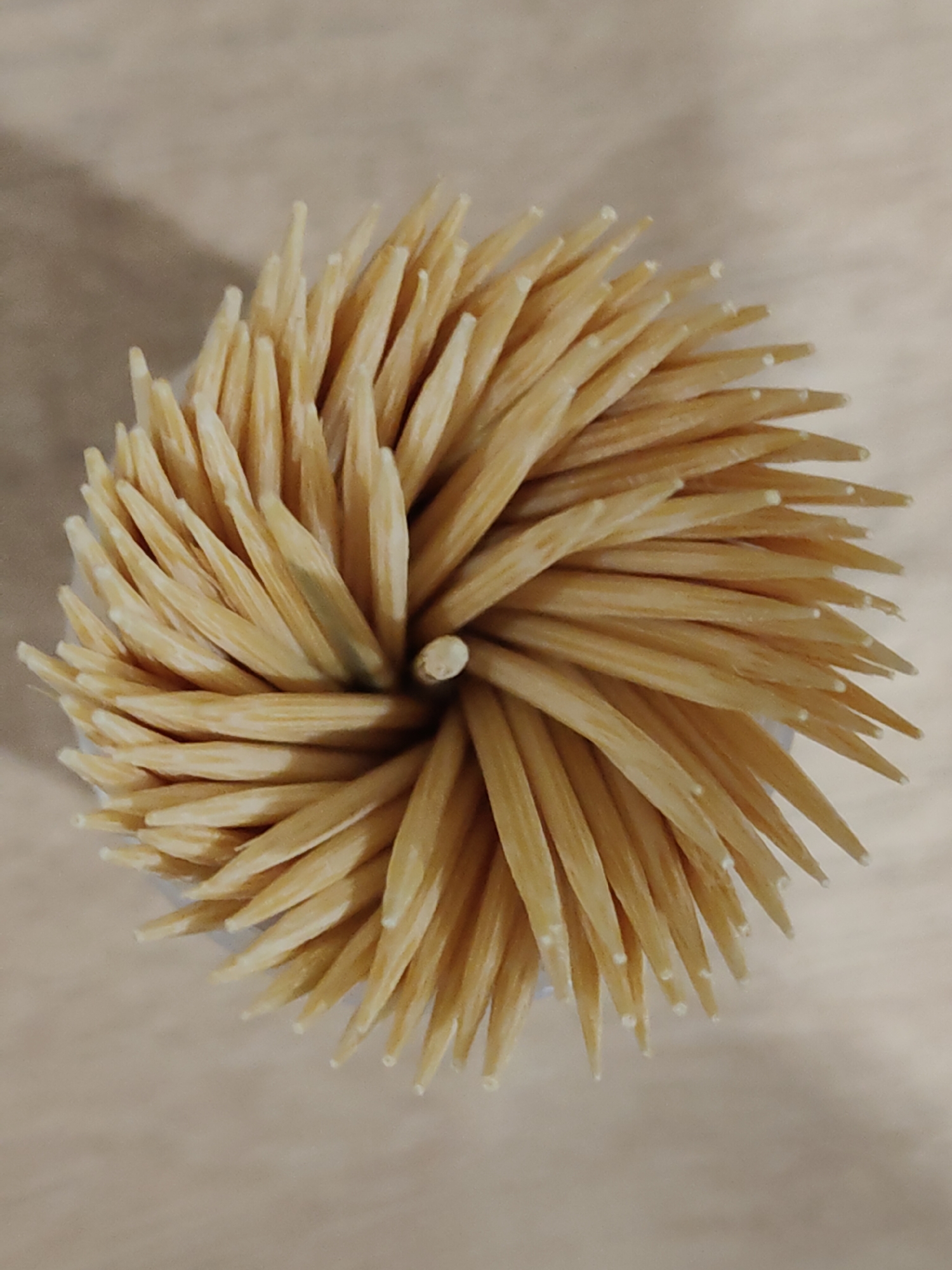 The image shows a close-up, top-down view of a collection of toothpicks arranged in a circular pattern. The toothpicks are made of wood, light tan in color, and tapered to a point. They are packed tightly together. The center of the circular arrangement is visible, revealing the cylindrical base that holds the toothpicks. The background is a neutral color, appearing to be a light beige or off-white surface. There is no indication of the location or city.