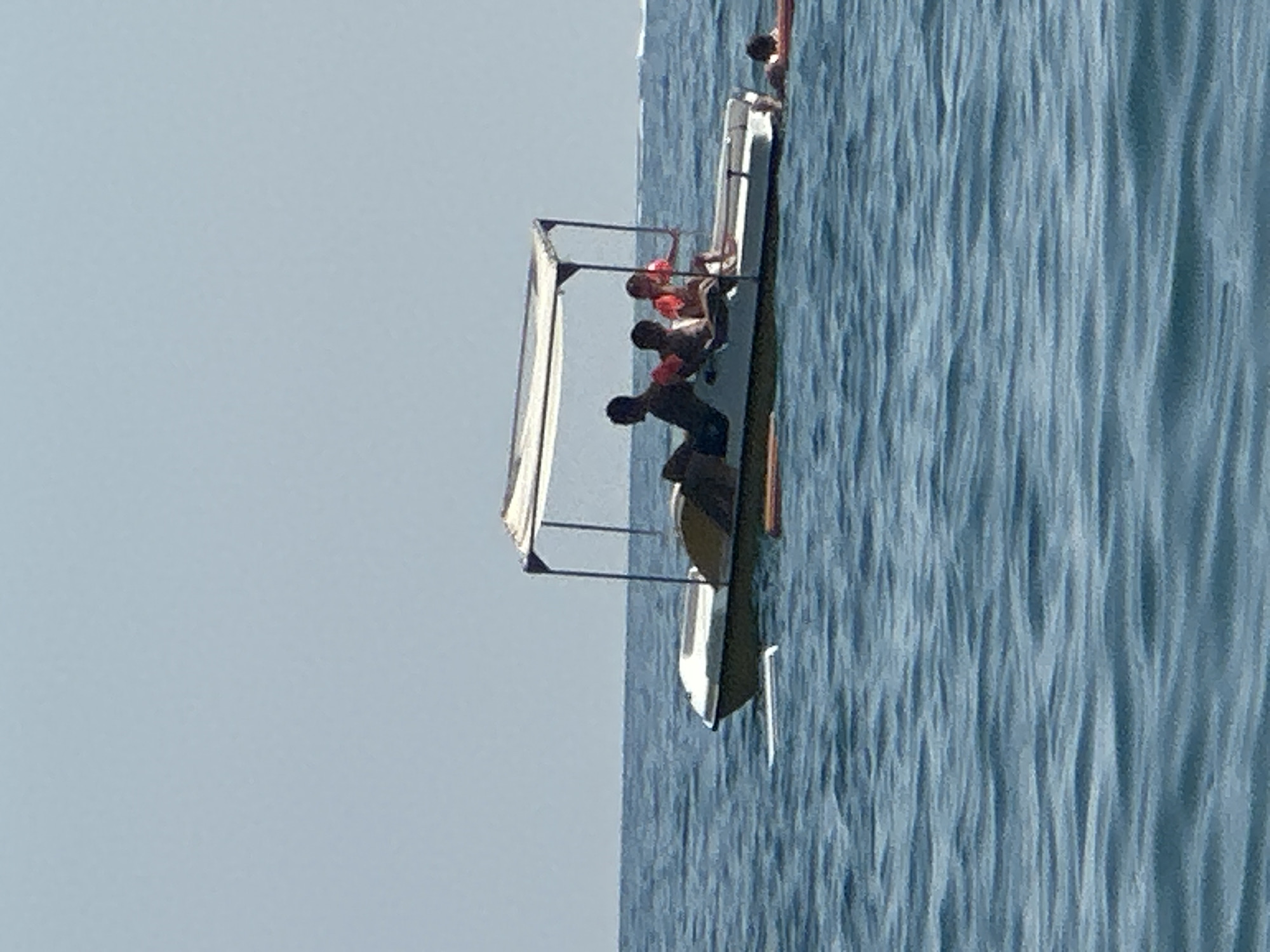 A pedal boat carrying four individuals is on the water under a clear sky. Two adults and two children are seated in the boat. The two children are wearing orange life vests. One person is partially submerged in the water to the right of the boat, wearing a life vest. The water is blue and has gentle ripples. The horizon is visible in the background, where the sky meets the water. The scene appears to be a leisurely outing on a body of water.
