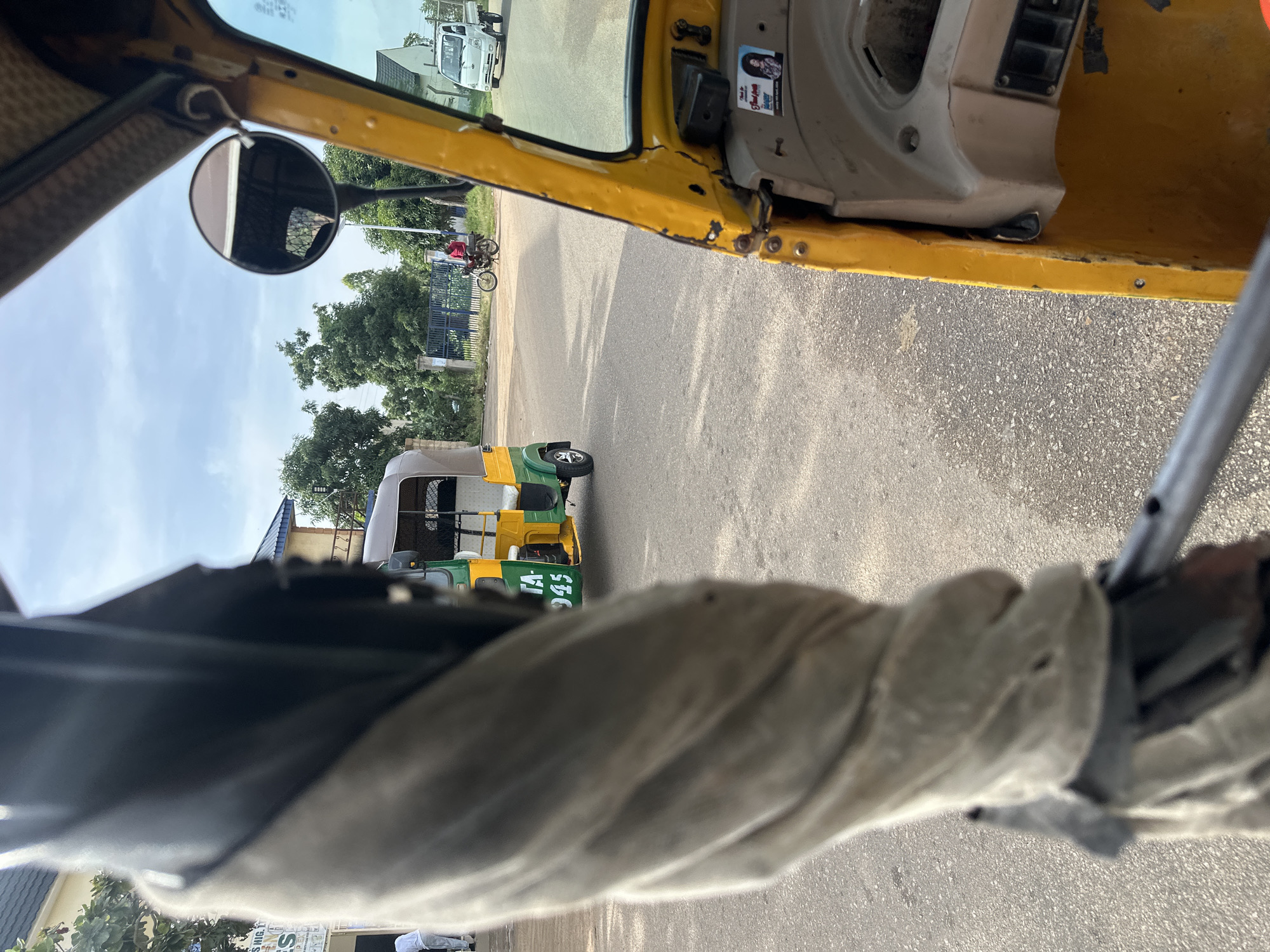 This is a first-person perspective image taken from inside a yellow auto-rickshaw, likely in Bukuru, Nigeria. In the foreground, a portion of the auto-rickshaw's interior, including a twisted fabric cover and part of the steering mechanism, is visible. The scene outside shows a paved road with an overcast sky. Several vehicles are present: another auto-rickshaw with "TA-945" painted on its side is in the middle ground, a white truck is visible in the distance on the right, and a person on a motorcycle is riding towards the left in the background. Trees and some buildings line the sides of the road. The overall environment suggests an urban or semi-urban setting with moderate traffic.