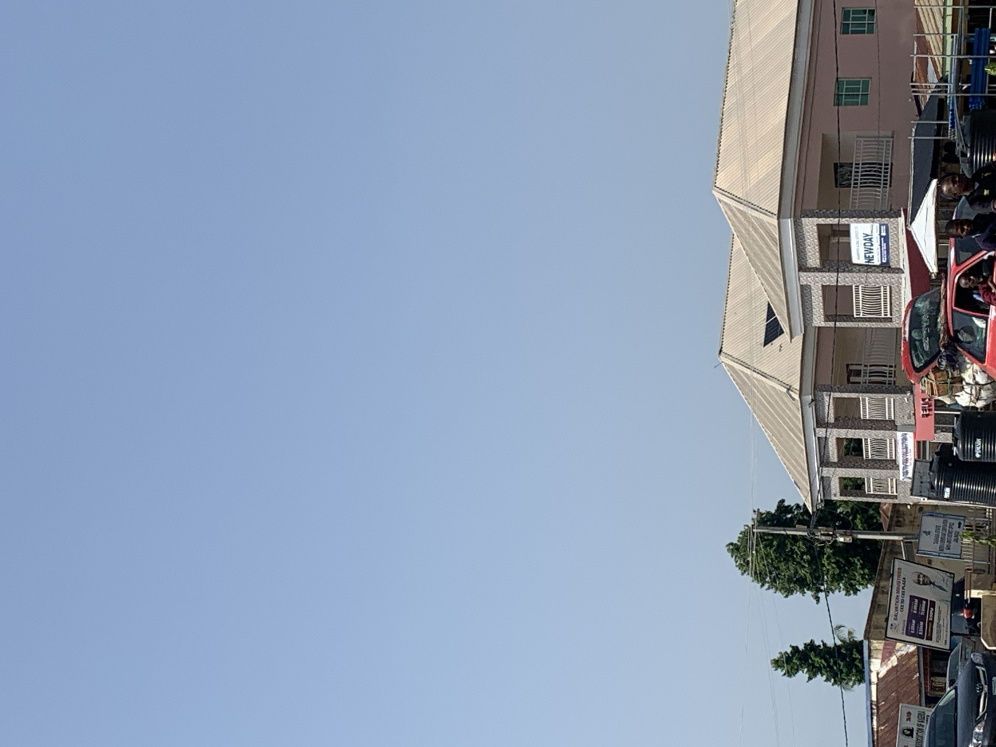 The image shows a street scene in front of a modern building with a light-colored, ribbed roof and pink walls. The building has multiple windows and balconies. A sign that reads "NEWDAY" is visible on the facade of the building. To the left of this building, there is another building with a corrugated metal roof and a sign that says "SALVATION MINISTRIES". In the foreground, there are two large black water tanks. A red car is parked in front of the building, and several people are visible around it. The sky is clear and blue. The environment appears to be an urban or semi-urban setting, possibly in Nigeria given the signage.