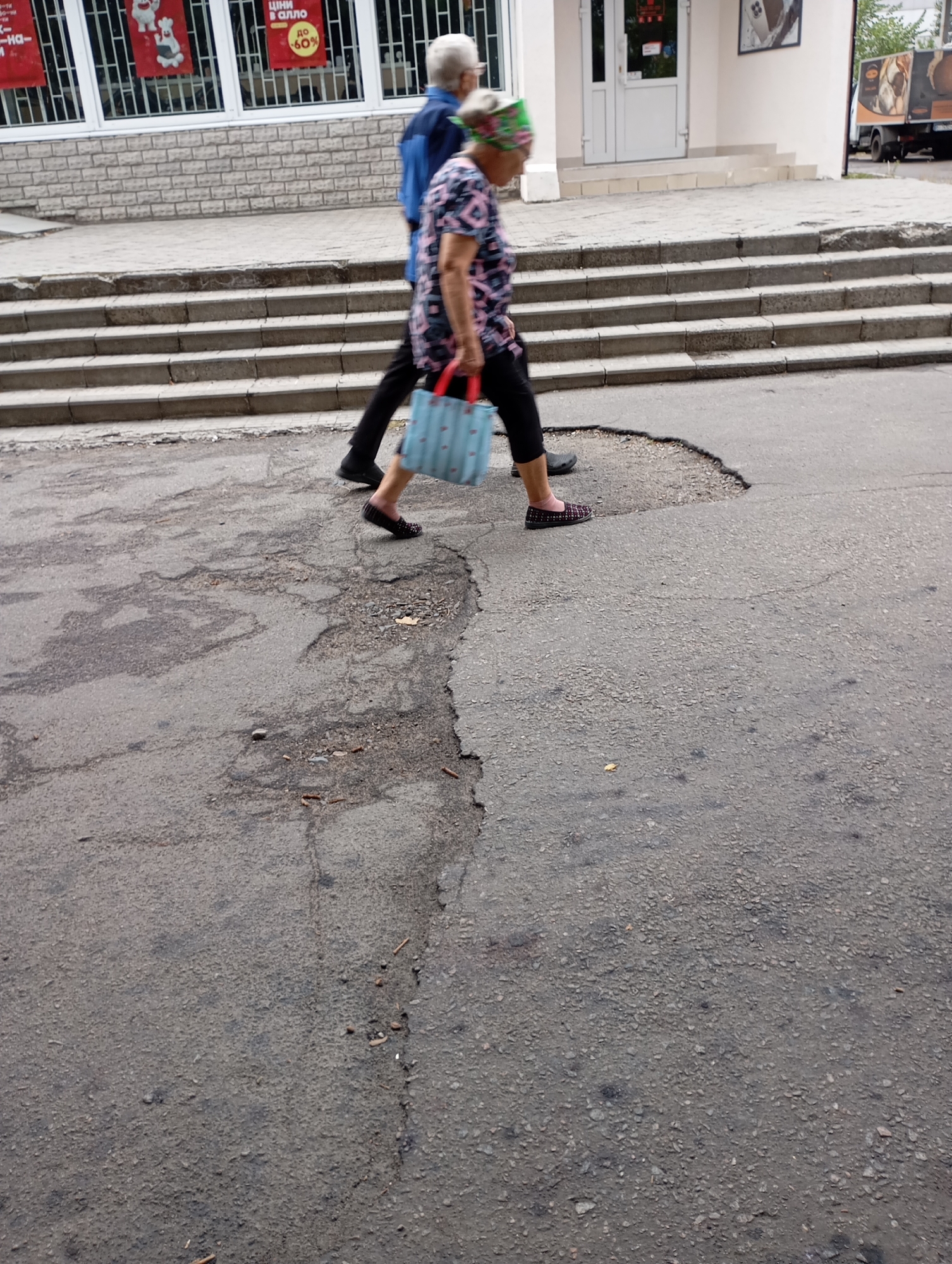 In Horishni Plavni, Ukraine, two individuals are captured mid-stride on an asphalt surface characterized by significant potholes and uneven wear. The person on the right, a woman, is wearing a colorful headscarf and a patterned dress, and carries a light blue tote bag with a repeating pattern of white hearts. She is also wearing dark, flat shoes. To her left, a man in a blue jacket and black pants walks alongside her. Behind them, a series of concrete steps leads up to a building. The building's facade features windows with red posters, suggesting a commercial establishment. The overall environment appears to be a public area, possibly a sidewalk or pedestrian walkway adjacent to a street.