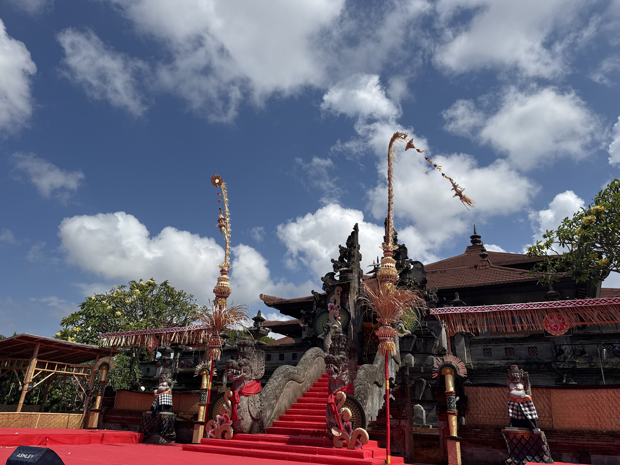 This is a low-angle, exterior shot of a Balinese temple or performance stage, likely in Denpasar, Indonesia, under a partly cloudy blue sky. The foreground is dominated by a bright red carpeted stage. To the left, a black speaker with "ASHLEY" printed on it is visible.
The main architectural structure in the midground is a temple-like building with traditional Balinese tiered roofs covered in dark tiles. The facade features intricate stone carvings, including two large, dragon-like statues flanking a bright red carpeted staircase. The staircase leads up to a platform in front of the building. There are decorative banners with red and white fringe hanging from the structure. Two tall, ornate poles with straw-like decorations and smaller hanging ornaments stand on either side of the staircase.
To the left of the main structure is a bamboo-covered open-air pavilion. Further to the right, a lush green tree with yellow flowers is visible.
The scene appears to be set for a performance or a cultural event, given the stage setup and decorative elements. There are no people visible in the image. The overall atmosphere is bright and festive, suggesting daytime. The image captures the vibrant colors and ornate details characteristic of Balinese architecture and culture.