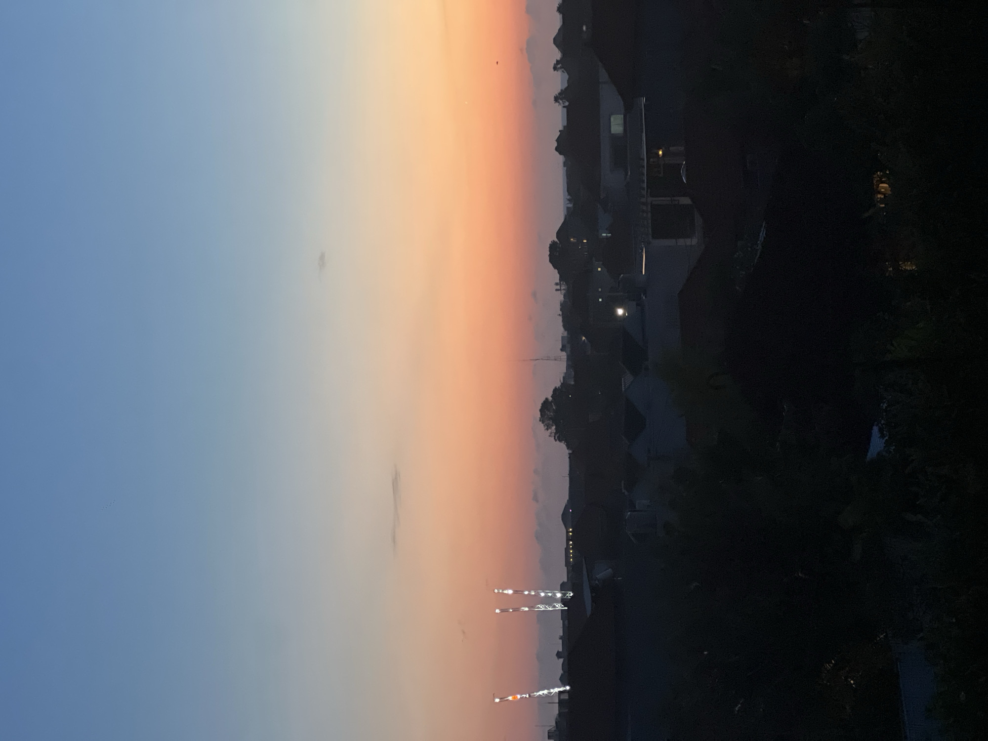 The image shows a sunset view over what appears to be a residential area. The sky transitions from blue to orange. The silhouettes of buildings and trees are visible against the colorful sky. In the lower portion of the image, there are lit-up construction cranes. Based on the prompt it is North Kuta, Indonesia.