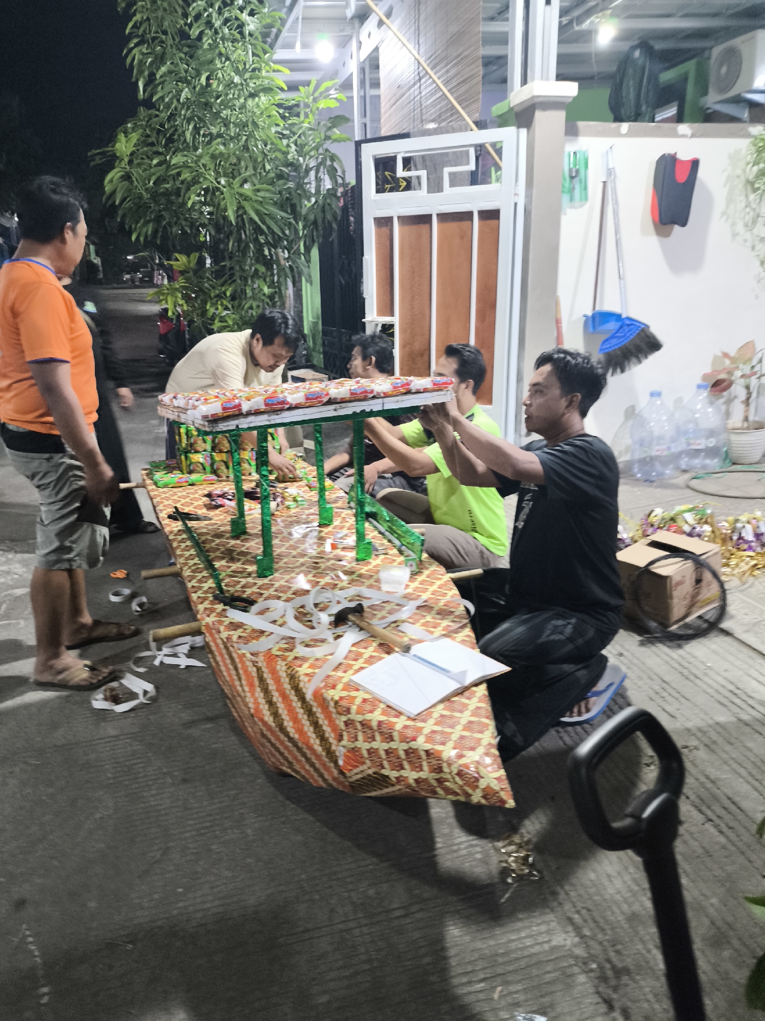 This is a medium shot, taken outdoors at night, likely on a street or driveway in Cilegon, Indonesia. The scene depicts several men engaged in preparing what appears to be a large, elaborately decorated structure.
In the foreground, on the right, a man in a black t-shirt and black pants kneels, holding a portion of the structure. To his left, a man in a bright green t-shirt and brown pants also participates in the assembly, his hands near the structure. Further to the left, a man in an orange t-shirt and grey shorts stands, observing or directing. Behind him, another man in a light-colored shirt is partially visible, also involved.
The central subject of the image is a long, rectangular structure supported by green legs, resembling a table or platform. It is covered with a patterned fabric, predominantly orange and brown, with white and yellow geometric designs. The structure itself is adorned with rows of packaged instant noodles. Surrounding the structure are various items: rolls of white tape, wooden tools, a notebook, and what appears to be decorative golden fringe. In the background, a white fence and a building with multiple doors are visible. Large plastic water bottles are stacked to the right, and a broom leans against the wall. A potted plant is also present in the background. The overall atmosphere suggests a community or family event, possibly a celebration or a religious observance, given the festive decoration.