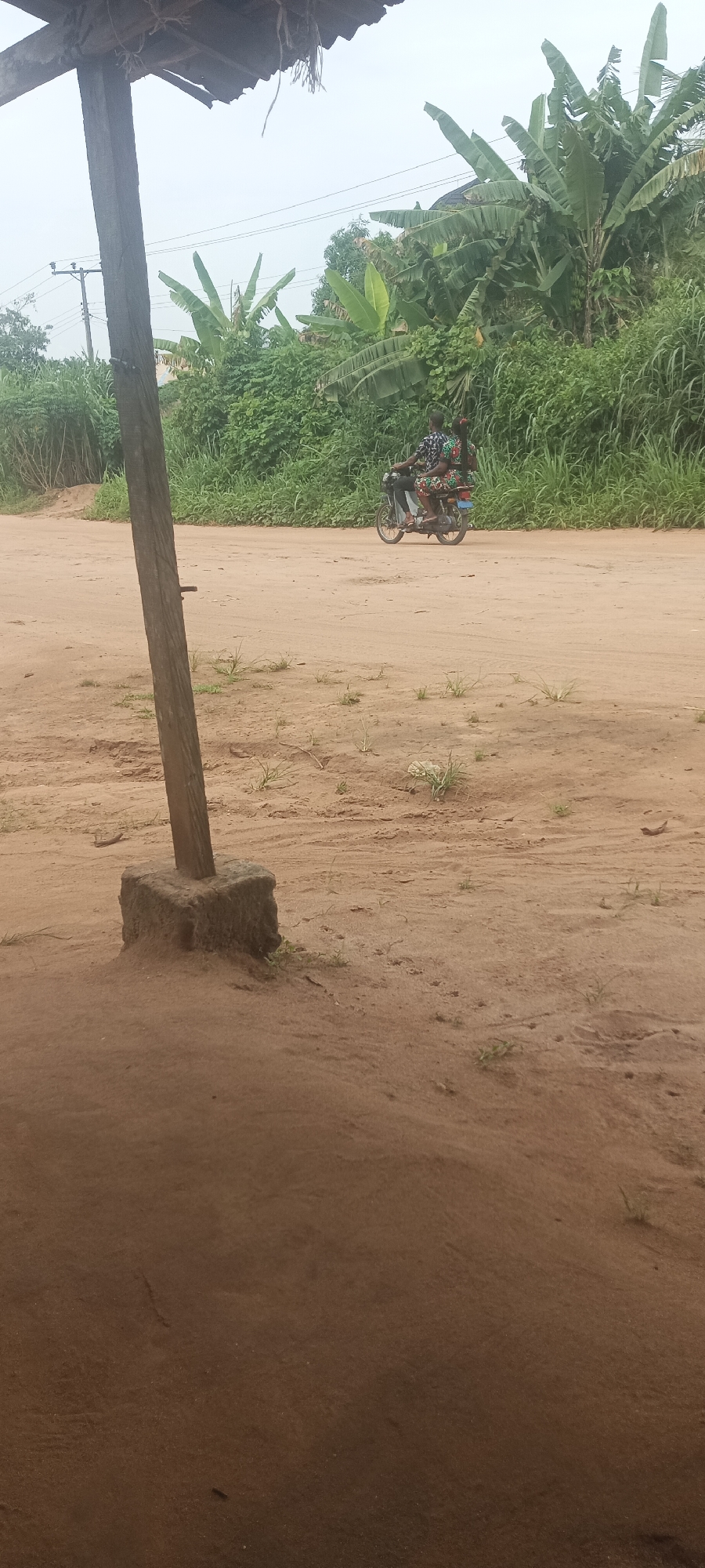 The image depicts a dusty, sandy landscape in Amauzu, Nigeria. In the mid-ground, a dirt road is visible, with a motorcycle carrying two people traveling along it. The road is flanked by dense green vegetation, including banana trees and other shrubs. In the foreground, a wooden post and its concrete base stand prominently, casting a shadow on the sandy ground. A simple roof structure is partially visible above the post. The overall scene suggests a rural or semi-rural setting.