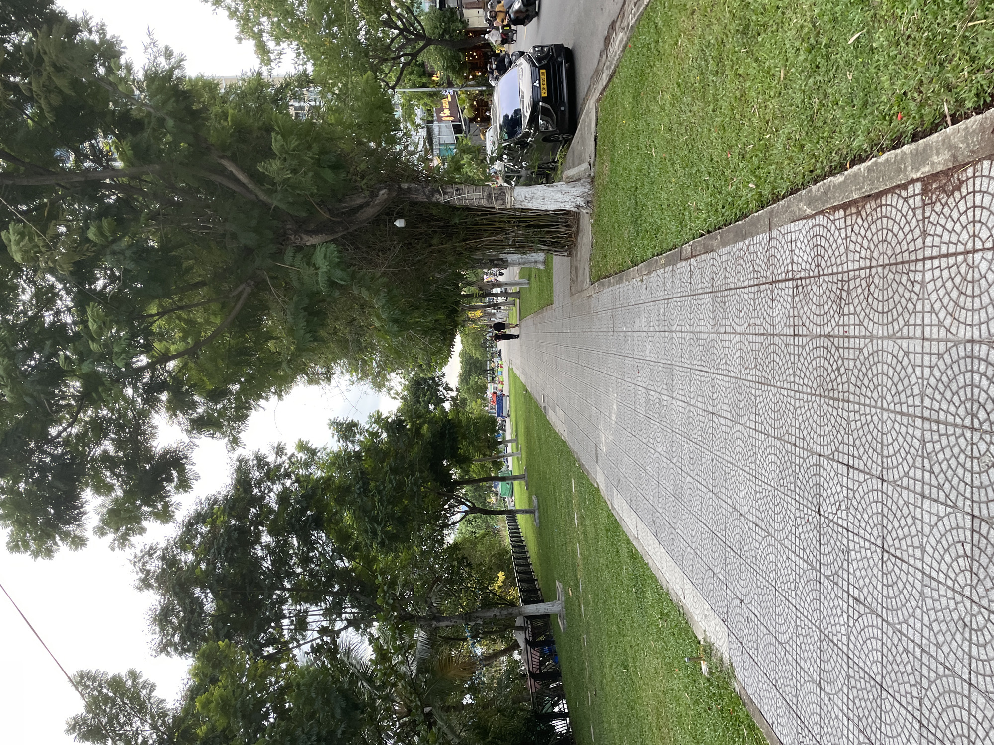 The image depicts a tree-lined sidewalk with a paved walkway in Hoi An, Vietnam. The sidewalk is made of decorative square tiles with a circular pattern. To the left of the sidewalk is a strip of grass, followed by a black metal fence and a body of water. On the right side of the sidewalk, there is a street with a black SUV parked on the side, followed by more grass and trees. In the distance, there are buildings and several motorcycles on the road. Two people are walking on the sidewalk in the middle ground. The sky is overcast.