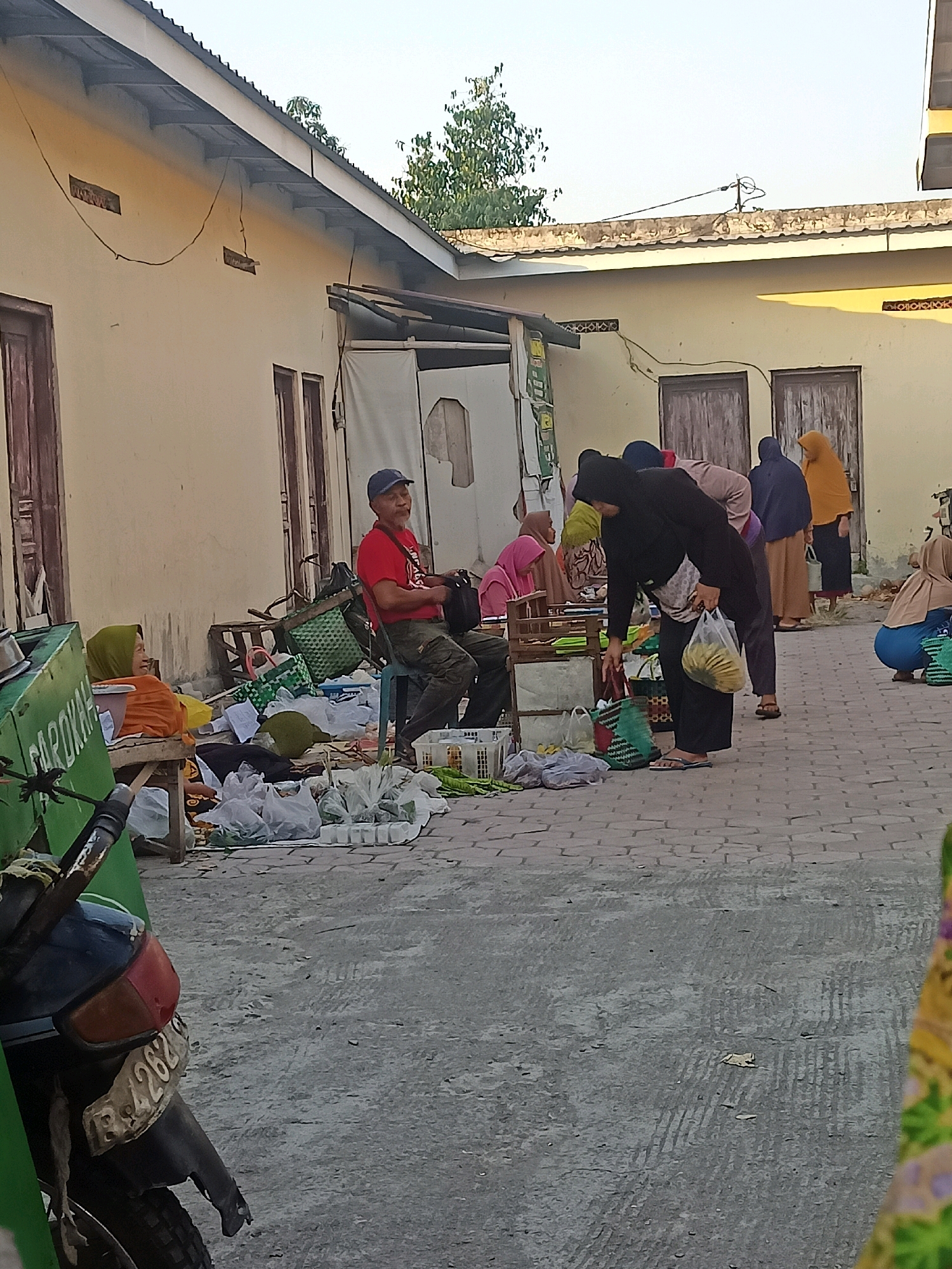 The image depicts an outdoor market scene, possibly in a courtyard, with several people engaged in activities related to selling or buying goods. In the foreground, a motorcycle is partially visible on the left, with a license plate displaying "B 4262 B". The ground is paved with hexagonal interlocking bricks. Several individuals are present. On the left, a woman in an orange headscarf and orange top is seated on a bench, looking to her left. Next to her is a green cart with text that is partially visible. In the center, a man wearing a red t-shirt, dark pants, and a blue baseball cap is seated on a blue chair, holding a black bag. In front of him are various items including bags of produce, a durian fruit, and woven baskets. Towards the right of the image, a group of people are gathered around stalls or displays. One person, dressed in black, is bending down and holding a plastic bag filled with bananas. Other individuals, mostly women wearing headscarves in various colors such as yellow, blue, and pink, are seen interacting or standing. The background shows a building with yellow walls, wooden doors, and a tin roof. The overall impression is of a bustling, informal marketplace during daylight hours, with a clear indication of commerce. The presence of the motorcycle with an Indonesian license plate strongly suggests the location is within Indonesia.