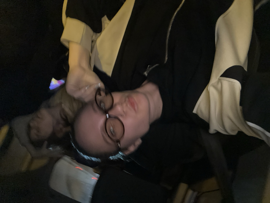 The image depicts two individuals within a dimly lit interior, located in Çiftlikköy, Turkey. In the immediate foreground, a female subject, with fair skin and dark hair pulled back from her face, directly observes the camera. She wears dark, oval-framed eyeglasses, a small hoop earring in her left ear, and a nostril piercing. Her lips are slightly pursed. She is dressed in a black top or jacket featuring a distinct cream or beige panel extending across the right shoulder and arm. A ring is visible on her right hand, which is positioned near her right temple. In the upper background, partially obscured behind the foreground subject, a second individual is visible. This person, consistent with female characteristics and lighter hair, is seen with their head tilted backward, indicating a state of repose. Their attire appears to be light-colored. The surrounding environment is characterized by profound darkness, with minimal discernible features. Vague, multi-colored light sources are evident in the upper-right quadrant, suggesting artificial illumination or reflections within a confined space.