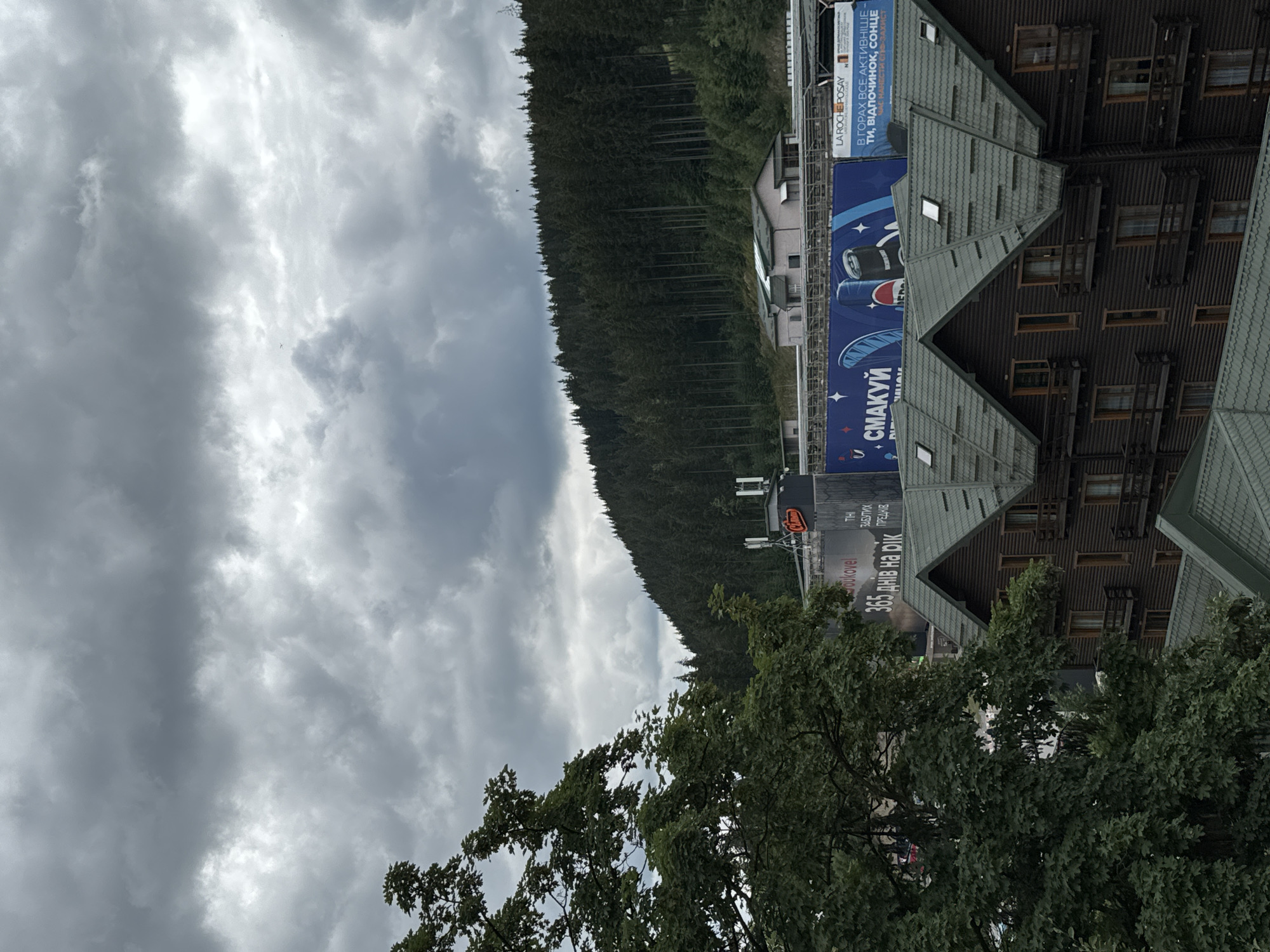 This media file captures a view of buildings nestled against a densely forested mountainside under a cloudy sky. The buildings appear to be multi-story with dark wooden exteriors and grey, sloped roofs. Some have visible windows and balconies. Large banners with text are attached to the side of the buildings, advertising products and services. One banner prominently features the brand "LA ROCHE-POSAY" and text in Ukrainian, including "В ГОРАХ ВСЕ АКТИВНІШЕ" (In the mountains, everything is more active). Another banner displays "365 днів на рік" (365 days a year) and a stylized image with the text "СМАКУЙ" (Taste). In the foreground, lush green trees with full foliage suggest a warm season. The overall impression is a resort or tourist area, likely in a mountainous region, given the text on the banners and the presence of the forest. The cloudy sky suggests overcast weather. No people are clearly visible in the image. The orientation of the image is rotated 90 degrees counter-clockwise.