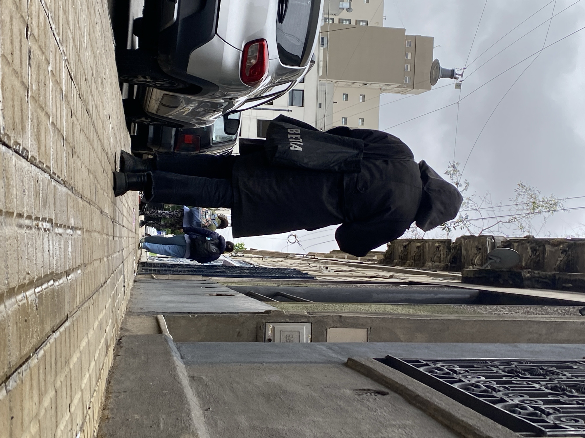 A man in a dark coat and pants walks down a narrow street in Buenos Aires, Argentina. Another person in jeans and a dark coat is walking in the same direction. A car is parked on the left side of the street, near a brick building. Across the street, there is a concrete building with ornate architectural details and a satellite dish. In the background, a tall building is visible under a cloudy sky. Electrical wires stretch across the sky.