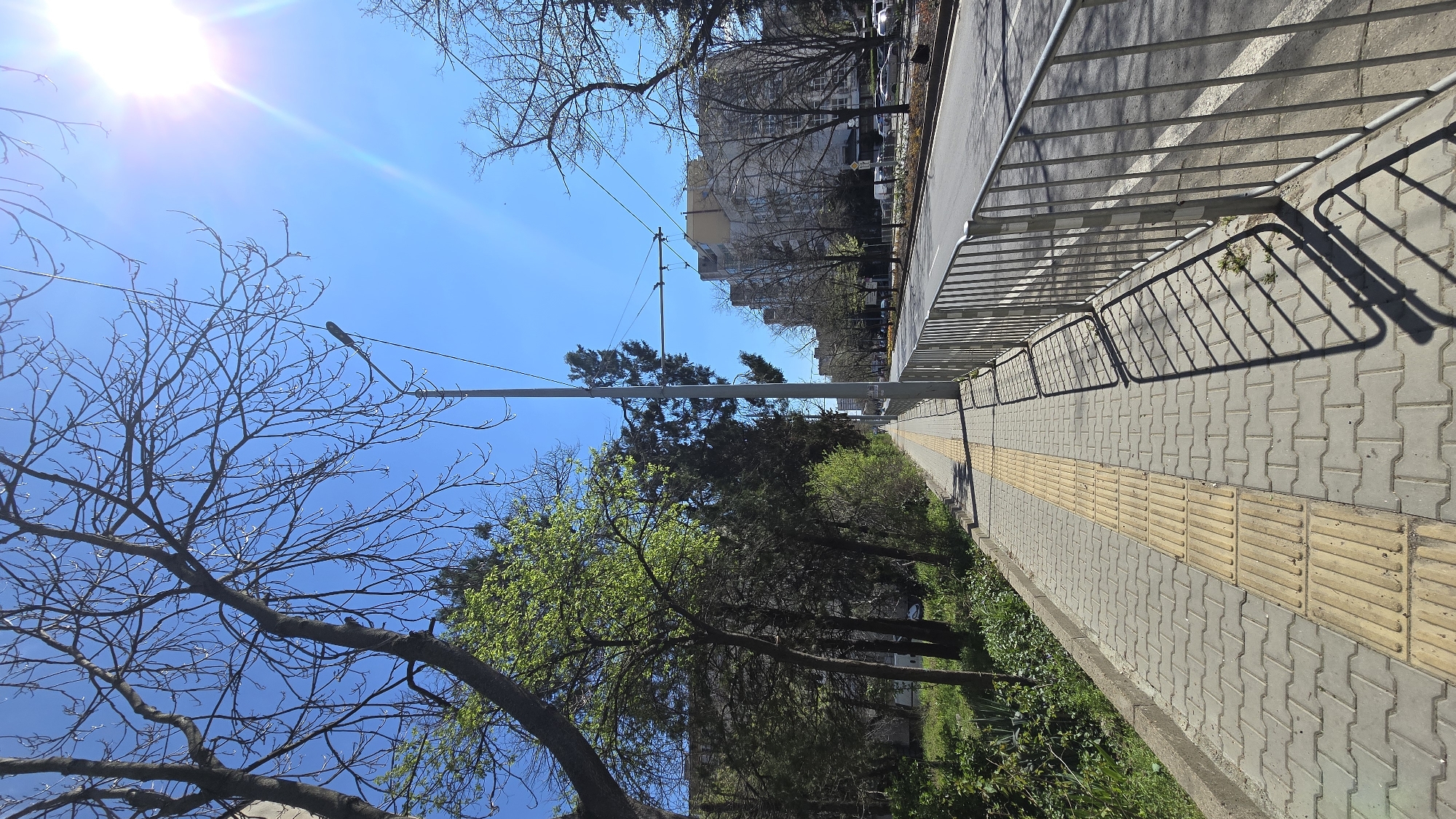 The image shows a pedestrian sidewalk in Stara Zagora, Bulgaria, flanked by a metal fence on one side and greenery on the other. A yellow tactile paving strip runs along the edge of the sidewalk. The street is visible beyond the fence, with buildings and trees lining it. Overhead, utility lines stretch across a bright blue sky with the sun shining. Bare branches and trees with new leaves fill the lower left part of the image.