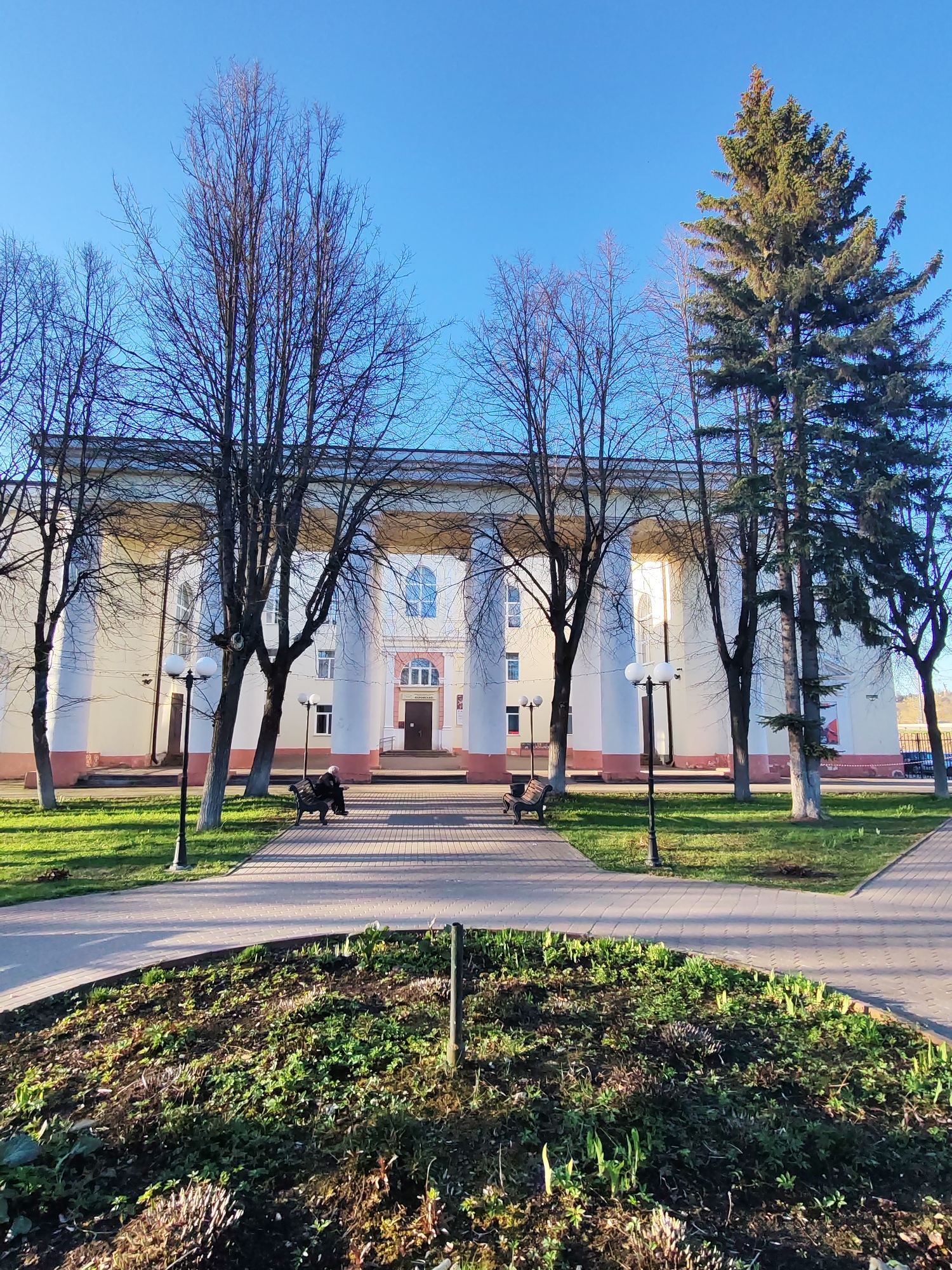A daytime, full outdoor shot depicts the House of Culture in the city of Yakhroma, Russia. A broad brick-paved walkway leads toward the building. The building has a row of white neoclassical columns capped with a gray band that forms the roofline of the colonnade. Windows are visible behind the columns, and a door is visible in the center of the building. The base of the building is painted a red hue. Trees, most of which are without leaves, frame the building on either side. To the right, a coniferous tree stands tall. A small, circular flowerbed is in the foreground filled with greenery and brown mulch. Lampposts line the walkway. Benches are situated along the walkway, and a person is seated on one. The sky is clear and blue.