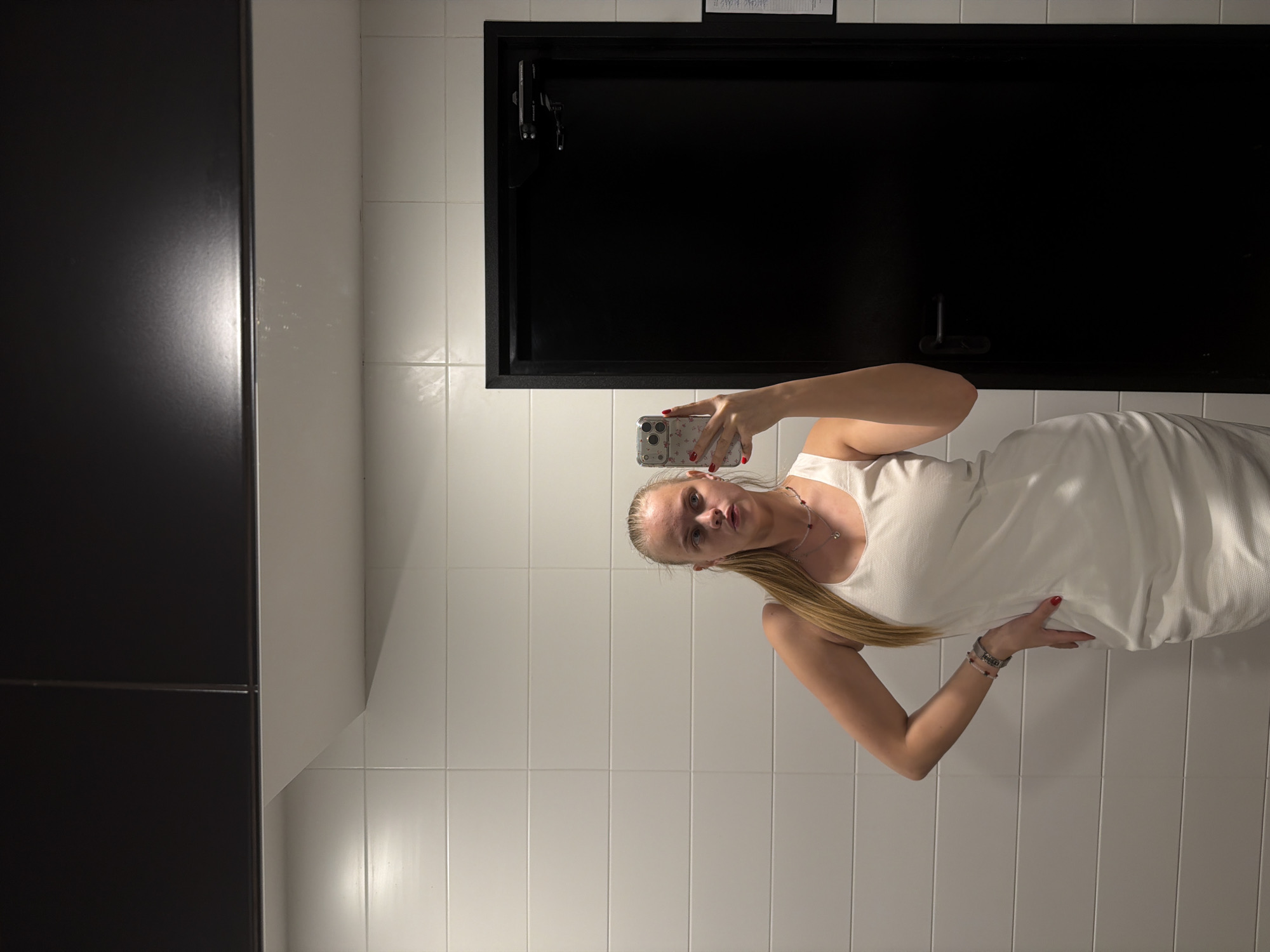 A woman with fair skin and blonde hair is taking a selfie in what appears to be a bathroom. She is wearing a white sleeveless dress and a necklace. Her nails are painted red. She is holding a smartphone with a patterned case. The background features white tiled walls and a black door with silver hardware. The scene is indoors, and the lighting appears to be artificial. The location is not possible to determine from the image.