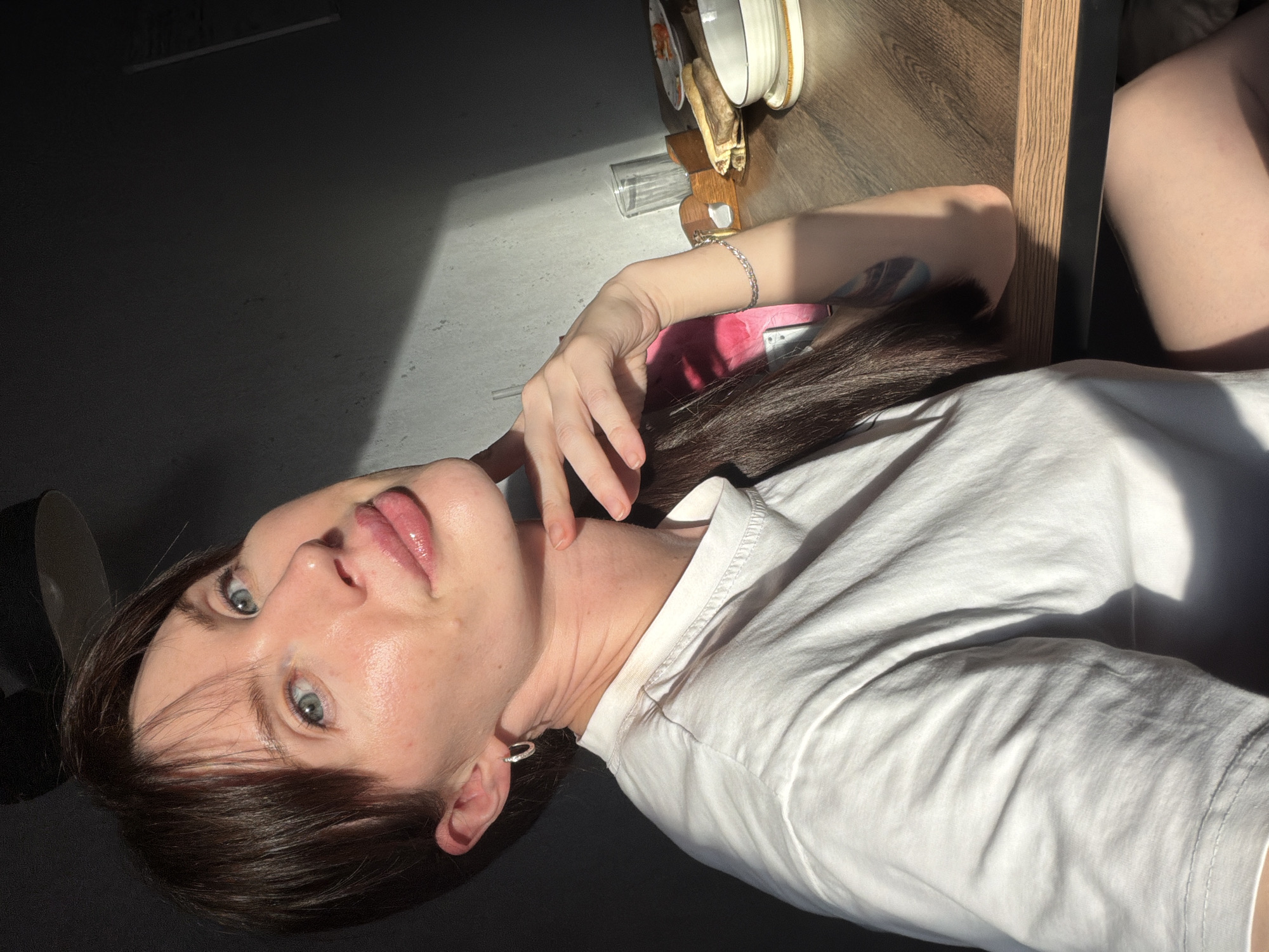 A selfie shows a woman in Perm, Russia, lying on her side and looking upward at the camera. She is wearing a white t-shirt and a silver earring in her left ear. Her hair is dark brown and partially covers her forehead. Her left hand is raised near her neck. The background features a table with dishes and a glass on it, a wall partially illuminated by sunlight, and a dark corner of the room.