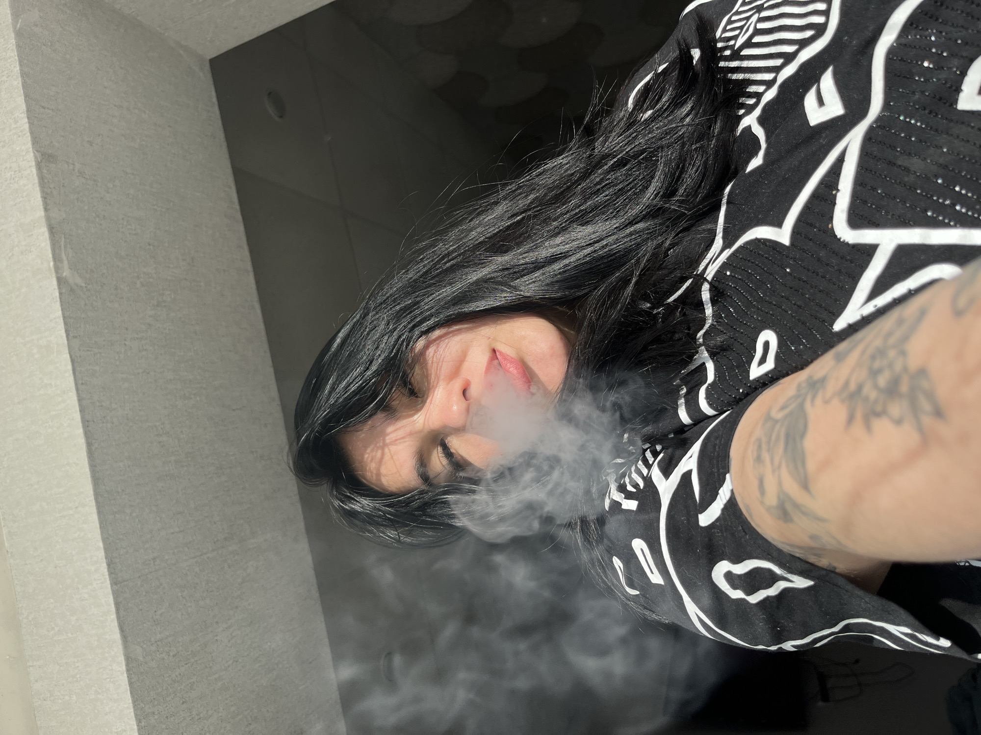 A person with long, black hair is exhaling a plume of smoke. The person is wearing a black and white patterned shirt. The person has a tattoo on their arm. The setting appears to be indoors, possibly in a room with a grey wall and a dark area in the background. There is no visible information to identify the exact location as Chaikovsky, Russia.