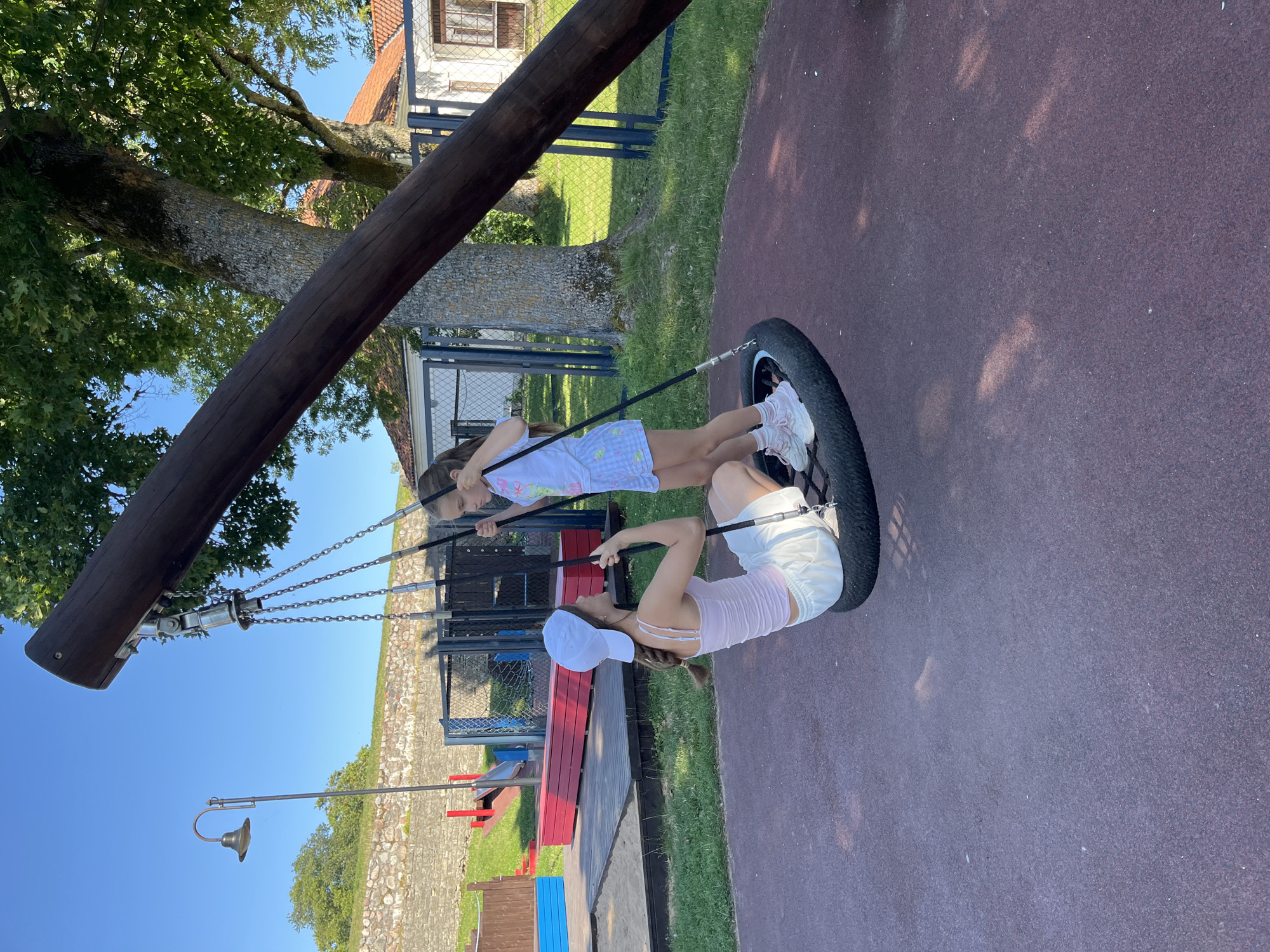 The image depicts a playground area with two young females on a tire swing. One female, wearing a white cap, light pink tank top, and white shorts, is seated on the swing, holding onto the chains. The other female, wearing a white t-shirt with a graphic and patterned shorts, is standing and appears to be interacting with the seated female. They are on a dark red rubberized surface, commonly found in playgrounds.
In the background, there is a large wooden beam supporting the tire swing, with metal chains connecting it to the tire. A mature tree with green foliage is to the right of the beam. Behind the playground equipment, there is a stone wall, a lamp post, and fencing. Further in the background, buildings with orange tiled roofs and white walls are visible. The sky is clear and blue, suggesting it is a sunny day.
This scene is consistent with the playground located at Kuressaare Castle in Kuressaare, Estonia. The castle's historical stone walls and the general architectural style of the surrounding buildings are recognizable features of this location.