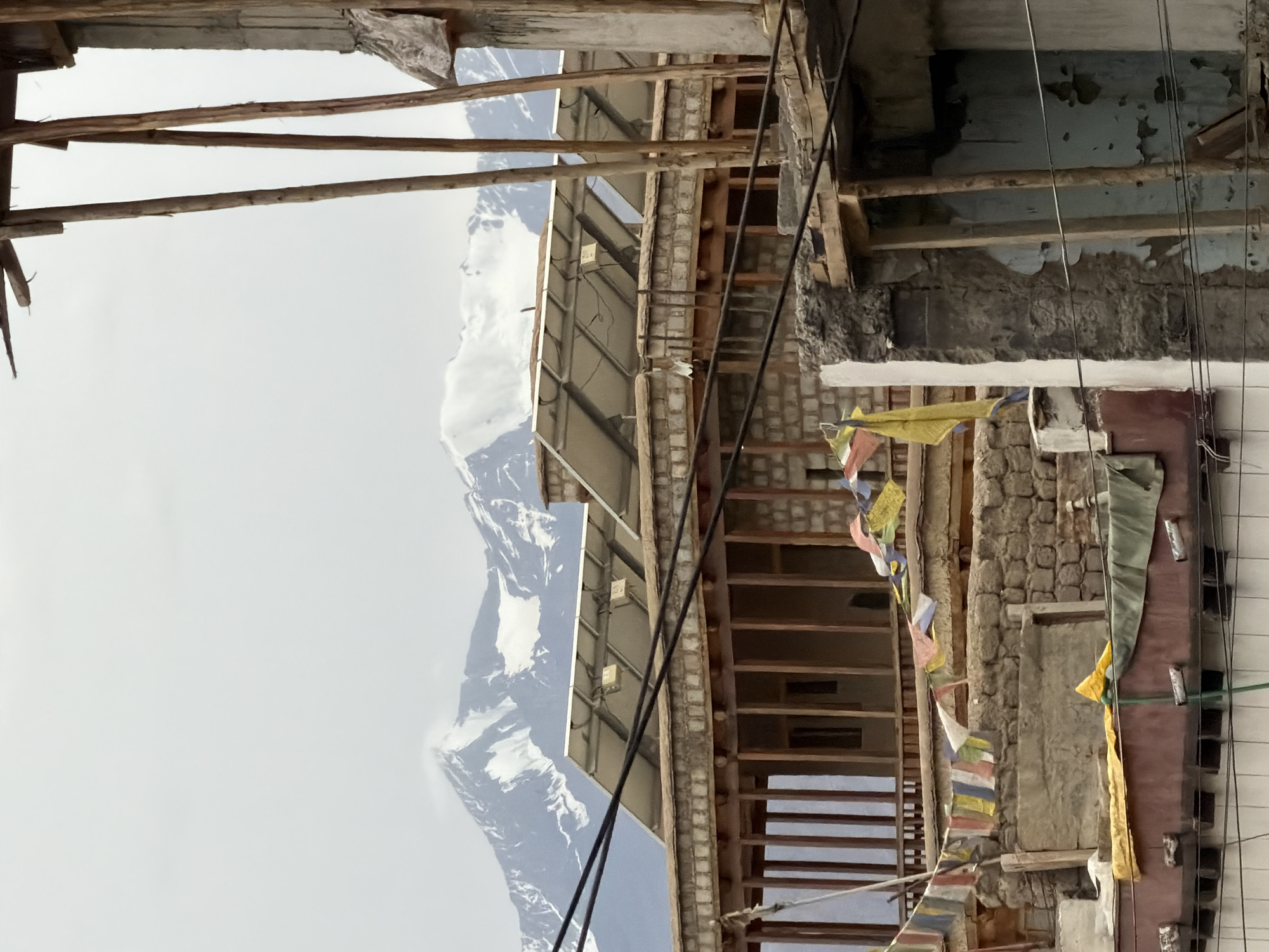 The image depicts a rooftop scene in Leh, India, featuring a building with solar panels installed on its roof. The building appears to be made of stone and wood, with a wooden balcony supported by pillars. Prayer flags are strung across the rooftop, adding a touch of cultural significance to the scene. In the background, snow-capped mountains rise majestically against a cloudy sky. The overall impression is one of a functional, yet culturally rich, urban environment nestled within a breathtaking natural landscape. The composition is dominated by the architectural elements of the building and the natural grandeur of the mountains, with the prayer flags serving as a vibrant accent.