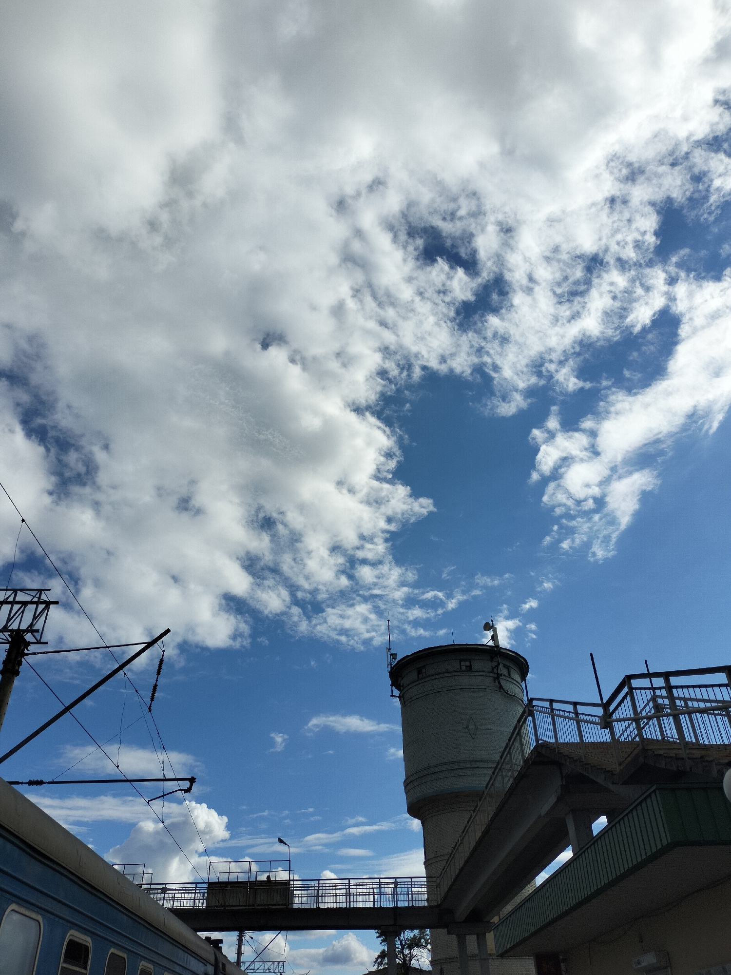 The image depicts a bright, daytime scene with a partly cloudy sky. In the lower left, a portion of a blue passenger train car is visible. Train tracks and overhead electrical lines are suspended by a metal support structure. To the right and slightly above, a light-colored, cylindrical water tower made of bricks stands next to an elevated walkway and stairs with metal railings. The walkway appears to connect to the water tower and extends over the train tracks. Below the walkway, there is a shaded area and a glimpse of greenery, possibly a tree. The city's location is not specified, as the information is unknown.
