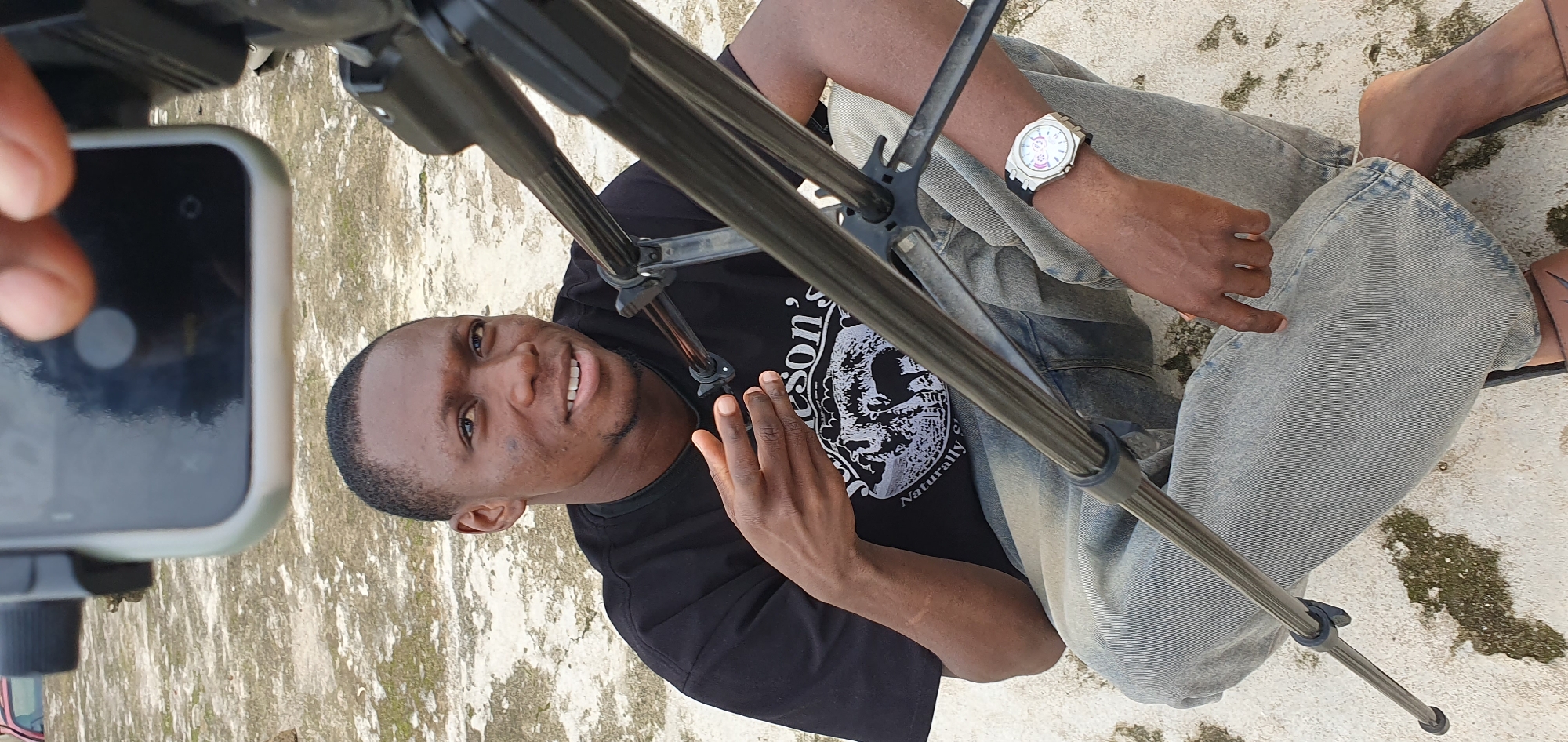 A young Black man is sitting on a concrete surface, facing slightly to his right and looking towards the camera with a gentle smile. He is wearing a black t-shirt with a graphic print featuring animals and text, and light-wash denim jeans. His left hand is raised, with fingers slightly spread, as if in the middle of speaking or gesturing. A tripod is positioned in front of him, with some of its legs visible. He is wearing a watch on his left wrist. The ground appears to be a light-colored, rough concrete surface, possibly outdoors. In the background, a portion of a red vehicle is visible. The overall scene suggests a casual, possibly creative setting, given the presence of the tripod, suggesting filming or photography. The location is stated to be in Anguldi, Nigeria.