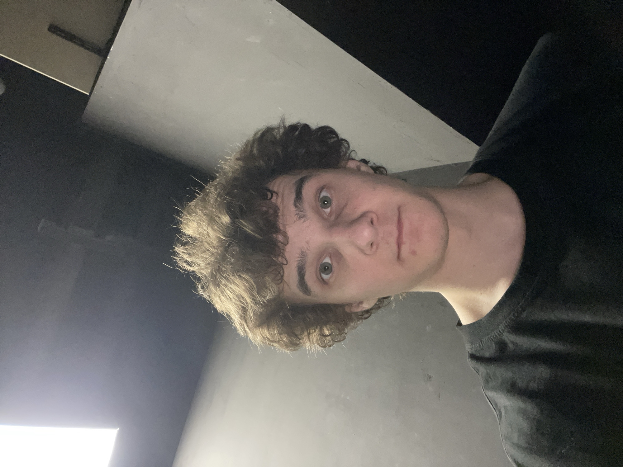 The image presents a close-up, tilted self-portrait of a young man, likely in his late teens or early twenties. He has fair skin, a pronounced jawline, and short, voluminous curly brown hair. He is wearing a dark, possibly black, crew-neck t-shirt. His gaze is directed straight at the camera, with his green or hazel eyes wide open, conveying a neutral or subtly surprised expression.
The background is minimalist and geometric, consisting of intersecting planes of dark grey and white or light grey walls and ceiling. Bright, artificial lighting illuminates parts of the background, creating a sharp contrast with areas shrouded in deep shadow. This suggests an indoor setting, possibly a hallway or a room with plain walls, under artificial light. The strong tilt of the camera gives the impression of the subject looking upwards or the environment being at an angle. There is no visible text, distinct objects, or specific architectural cues to definitively identify the location as Krasnoyarsk, Russia, beyond the context provided. The time of day is not discernible due to the indoor lighting.