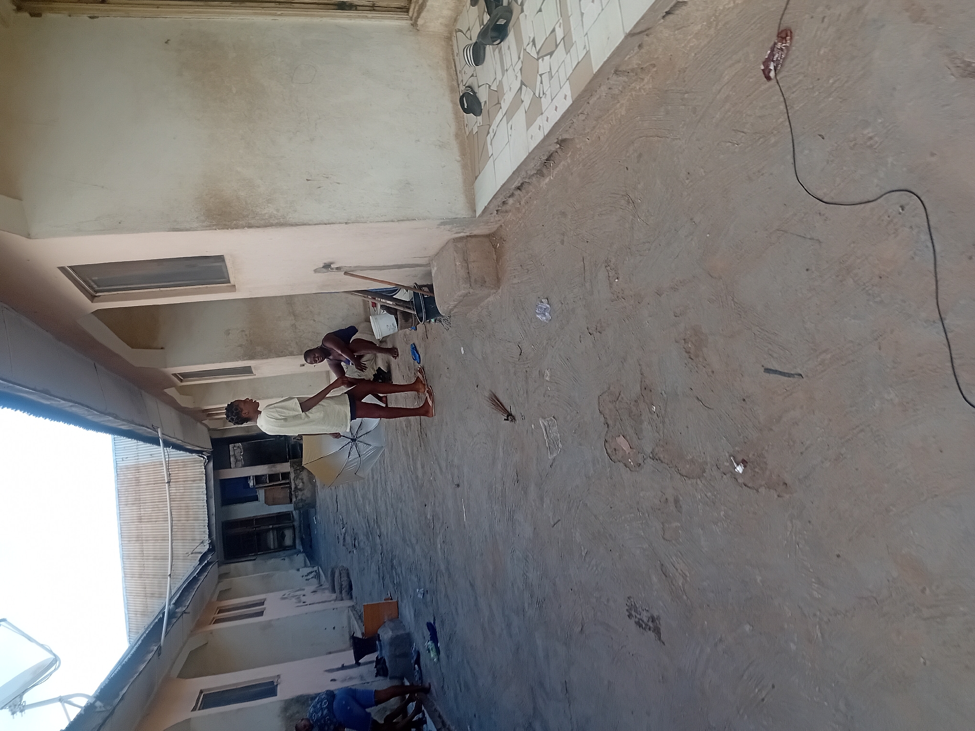 This image depicts a communal outdoor or semi-outdoor space, likely a courtyard or passage between residential buildings, characteristic of Lokoja, Nigeria. The scene unfolds under bright daylight, suggesting a clear sky.
Several individuals are present. In the central left, a person wearing a light-colored t-shirt and dark bottoms stands, holding a white or cream-colored umbrella. Further back and to the right, another person in a dark top and shorts is kneeling or squatting near a white bucket, seemingly engaged in a chore. In the lower left, a person in a patterned blue garment is partially visible, sitting or squatting. A fourth individual is partially visible in the upper right.
The ground is a worn, unpolished concrete slab, showing signs of use and dirt. Near the kneeling individual, several pairs of sandals or flip-flops are arranged beside a wall section adorned with geometric brown and beige tiles. A long black electrical cable extends across the concrete floor. The surrounding structures feature simple, light-colored walls, some with visible stains and wear. Rectangular windows and doorways lead into various rooms. In the background to the left, a white satellite dish is visible, along with corrugated metal roofing.
The activity suggests everyday life in a modest, communal setting. The bright lighting indicates it is daytime, likely midday or afternoon. No discernible text is visible in the image.
