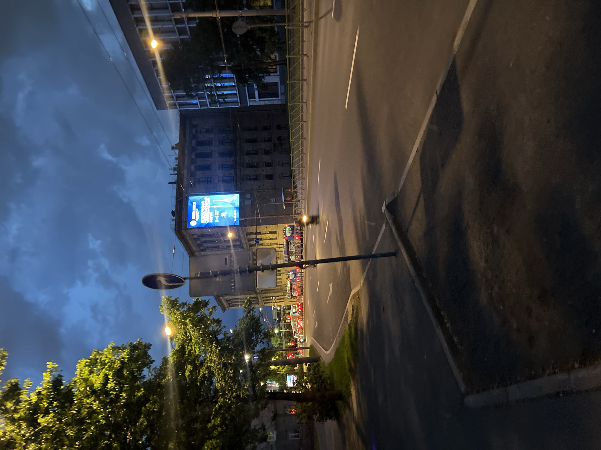 This image captures an urban street scene at dusk or early night in Saint Petersburg, Russia. The sky above is dark blue-grey with distinct cloud formations, indicating an overcast condition.
In the immediate foreground, a dark asphalt pavement with a grey concrete curb is visible on the left, leading to a darker, unpaved area. Centrally positioned in the lower mid-ground is a dark metal pole supporting a circular blue regulatory sign featuring a white arrow pointing diagonally right and upwards.
To the left of the road, a sidewalk is lined with mature trees featuring bright green foliage, illuminated by artificial light sources, likely streetlights, which also cast yellow-orange glows onto the scene. The main thoroughfare is a multi-lane asphalt road, appearing damp, with clear white lane markings. In the middle distance, a row of red traffic lights is active, indicating that traffic is stopped. Several vehicles, primarily identified as modern buses or trams with illuminated blue interiors, are halted on the road. A single motorcyclist, rendered with slight motion blur, is visible on the road moving rightward. A low, light-colored metal fence separates the road from a narrow strip of grass.
Architecturally, on the left side of the street, an older, multi-story building with a dark, classical facade and multiple windows is present. Further into the mid-ground, another substantial building of an older architectural style hosts a prominent, brightly illuminated digital billboard on its facade. The billboard displays text in Cyrillic, clearly showing "МЕДИА ФОРУМ" (Media Forum), "ДНИ РАЗВИТИЯ СЕВЕРО-ЗАПАДА" (Days of Development of the North-West), and "С-ПЕТЕРБУРГ 3-6 ИЮНЯ" (St. Petersburg June 3-6), confirming the location and event. To the right of this building, a more contemporary, multi-story building with a lighter facade and numerous large, rectangular windows, some reflecting a blue light, completes the street-front on that side. The scene is illuminated by multiple artificial light sources, including streetlights and vehicle lights, contributing to the varied light levels across the image. No individuals are clearly discernible outside of vehicles.