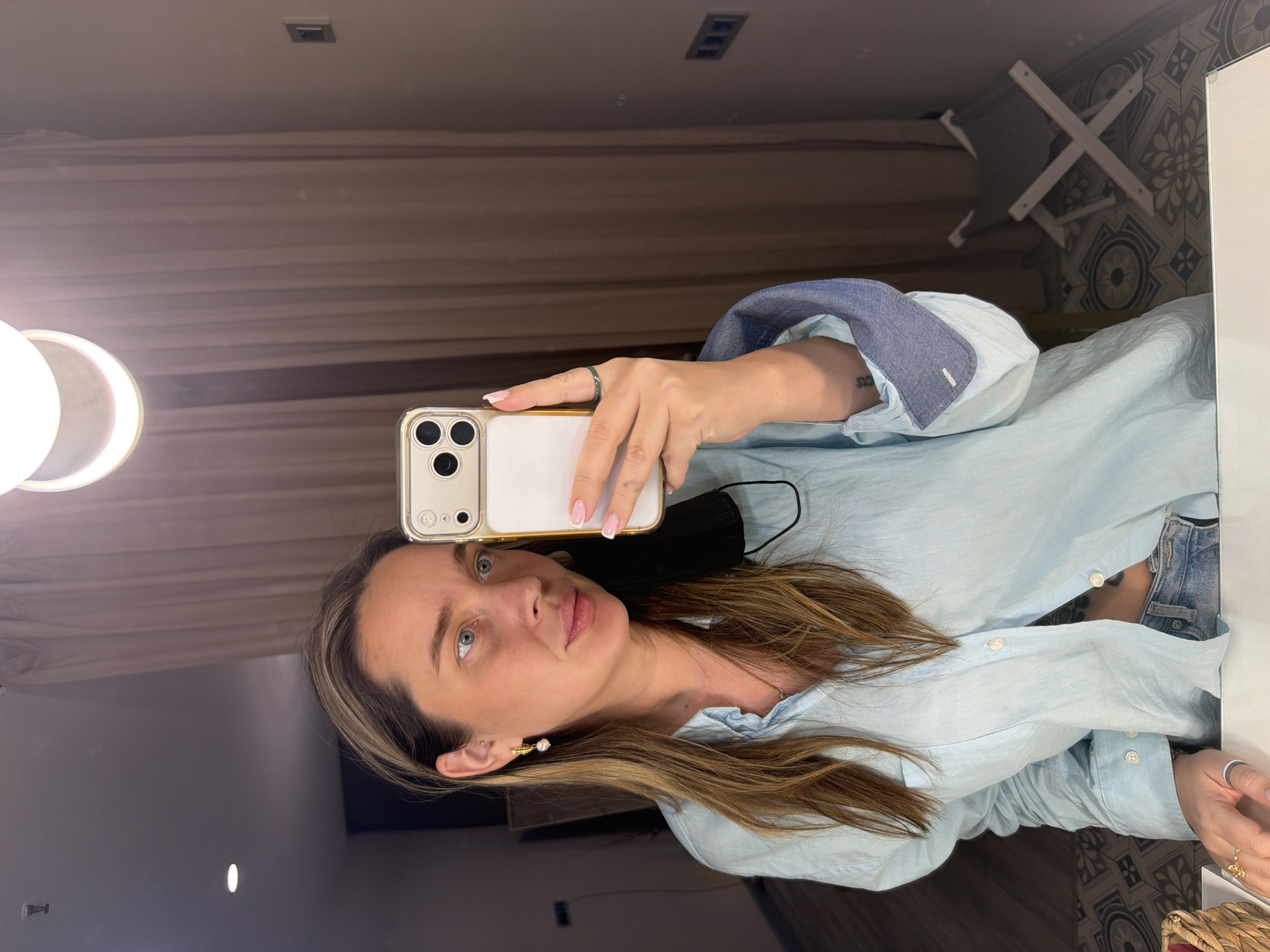A woman with long brown hair, wearing a light blue button-down shirt and pearl earrings, is taking a selfie in a mirror. She is holding a gold-colored iPhone with three lenses, partially obscuring her face. A black face mask is hanging around her neck. Her nails are neatly manicured with a white tip design. A green ring is visible on her finger. In the background, a ring light is mounted on the wall, and a clothes drying rack is standing near patterned wallpaper. The location is likely indoors, possibly a dressing room or bathroom. There is no evidence to suggest the location is in Moscow, Russia.