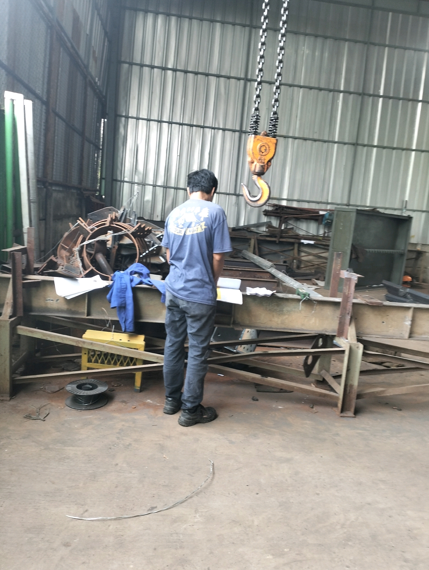 In an industrial workshop located in Gunung Sindur, Indonesia, a man stands with his back to the camera, wearing a blue t-shirt and dark pants. He appears to be looking at a stack of papers on a yellow industrial stool. The workshop is filled with various metal structures, scrap metal, and construction materials. A large industrial crane hook is suspended from the ceiling. The floor is concrete, and the walls are corrugated metal. The overall scene suggests an active but somewhat disorganized workspace.