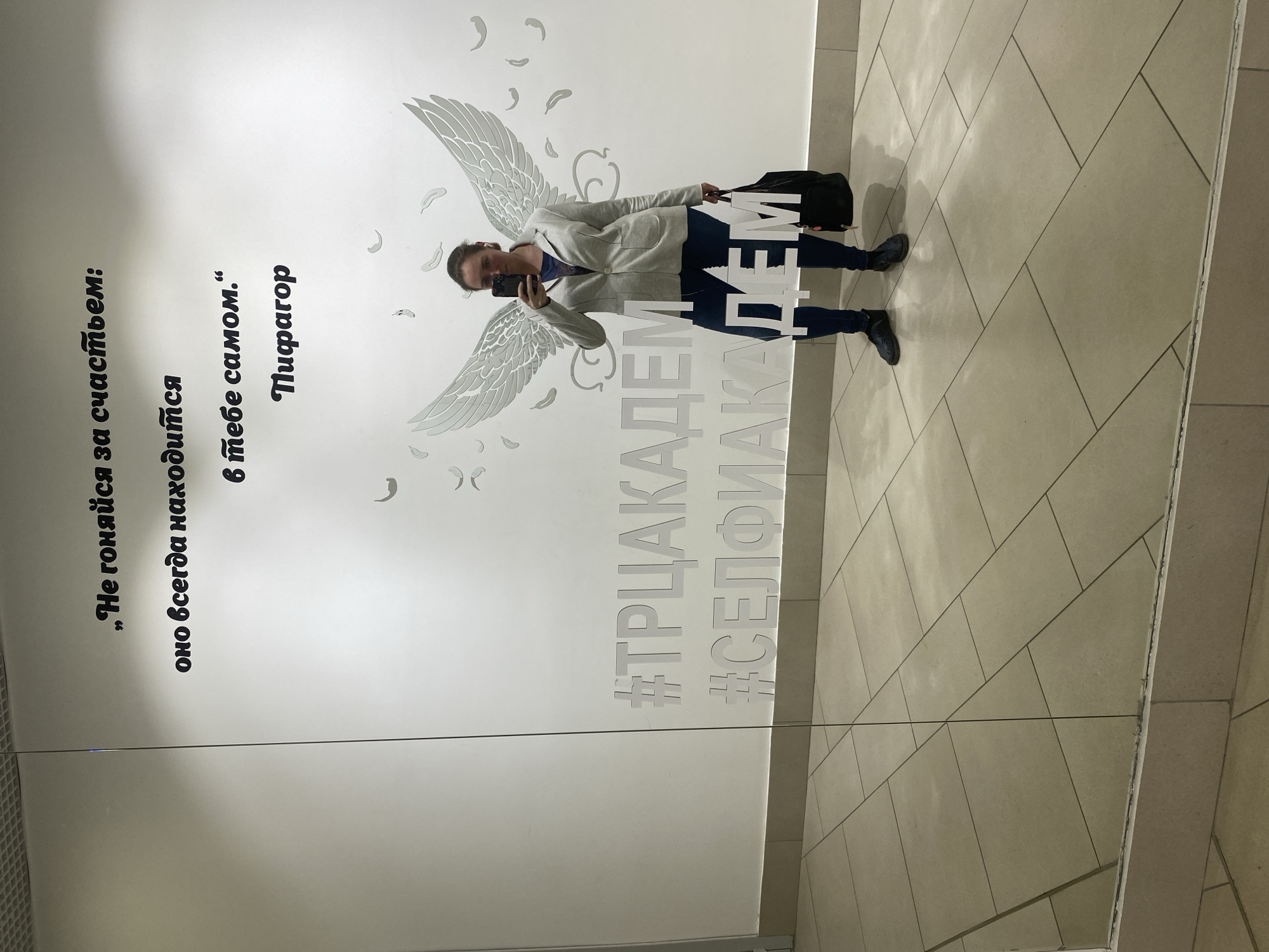 Here is a detailed description of the image:
**Scene:** An individual takes a selfie in front of a promotional wall inside what appears to be a shopping center. The location is identified as Yekaterinburg, Russia based on the Cyrillic text on the wall. The wall has text and decorative wing designs on it. The image is slightly rotated, with the person appearing off-balance.
**People:**
* One person, who appears to be female, holding a smartphone and taking a selfie. She is wearing a light gray blazer, dark blue jeans, and dark shoes. She is also holding a dark-colored bag in her other hand.
**Objects:**
* Smartphone: Used for taking the selfie.
* Bag: A black or dark-colored bag held by the person.
* Wall decorations: The wall features stylized angel wings.
* Text on the Wall: Cyrillic text is visible including a quote by Pythagoras and hashtags that contain the word "Academy" likely referring to the shopping center.
* Tiled Floor
**Actions:**
* Taking a selfie: The main action is the person taking a picture of themselves.
* Posing: The person is posed in front of the wings
**Environment:**
* The setting is indoors. The floor is tiled. The environment is clean and modern.