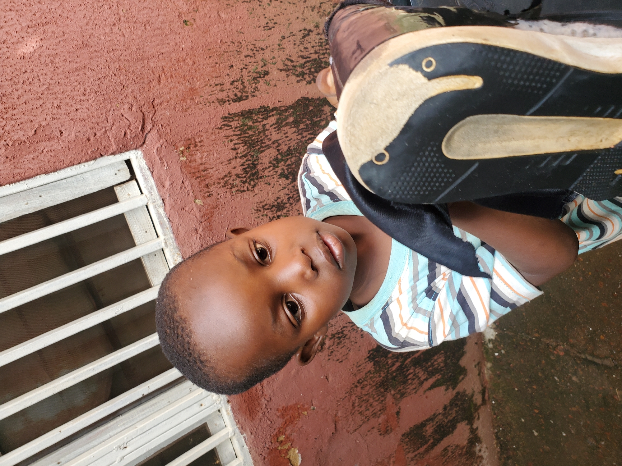 This is a medium close-up, portrait-style photo of a young boy, likely of African descent, lying on his back outdoors. He is wearing a striped t-shirt with thin blue, white, orange, and black stripes, and a black garment, possibly part of a shirt or jacket, is visible around his shoulders. His face is the primary focus, with his dark eyes looking slightly upwards. His skin is smooth and dark, and he has short, dark hair.
Behind him, a textured, reddish-brown wall with some patchy green moss is visible. To his left, a white slatted window or grate runs along the wall. Part of a black and tan shoe is prominently featured in the foreground, obscuring the lower half of his body and appearing to be placed over his torso. The lighting suggests it is daytime, and the presence of moss on the wall hints at recent rain or a humid environment. The overall scene feels casual and candid, possibly taken in a residential area in Jos, Nigeria, given the provided location context. There is no visible text in the image.