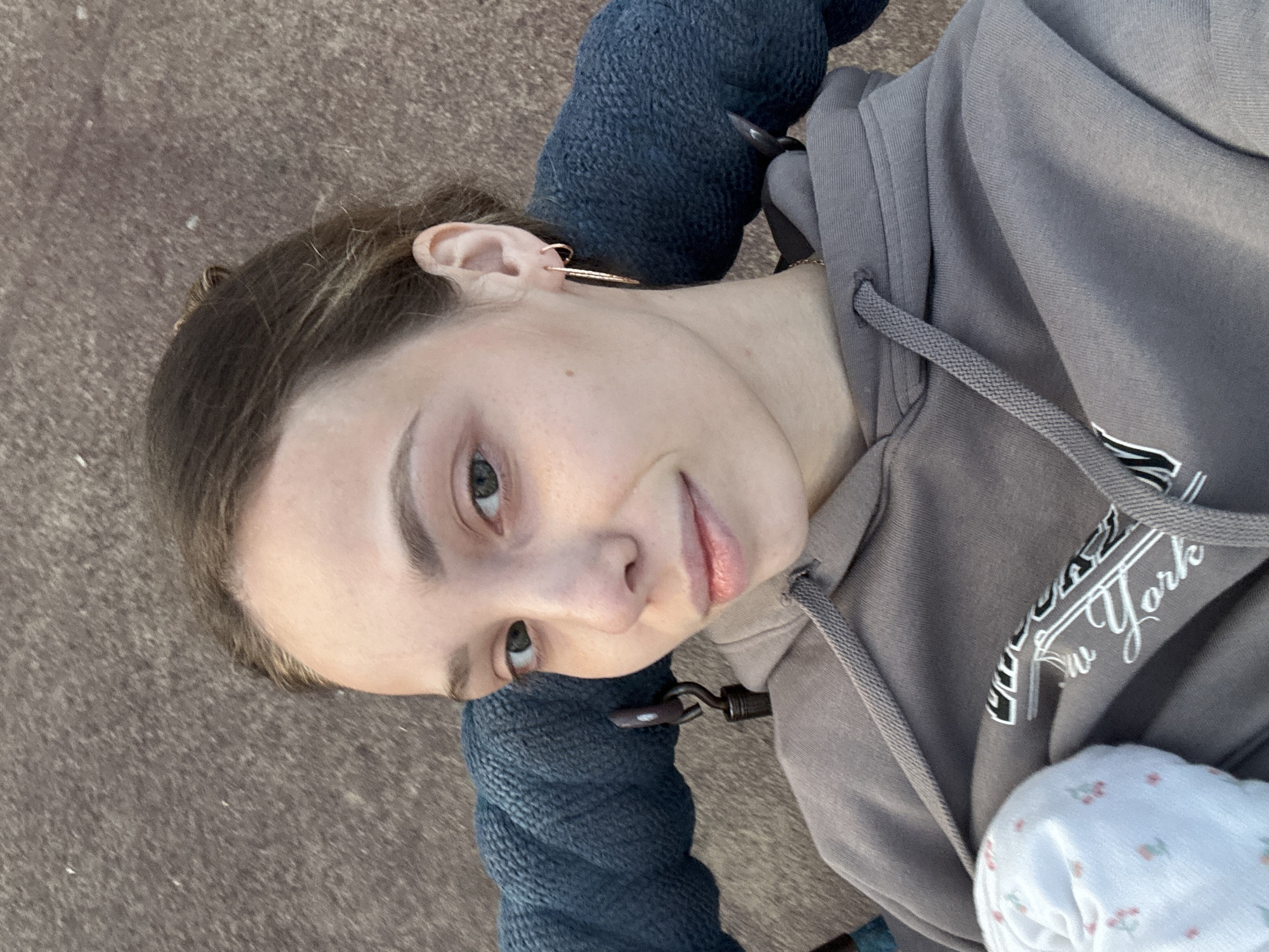A young woman with light brown hair pulled back is looking at the camera in a selfie. She is wearing a grey hooded sweatshirt and a gold hoop earring in her right ear. Her left arm is obscured by a thick, dark blue knitted fabric, possibly part of a chair or blanket. A small portion of a white baby blanket with a floral pattern is visible on her left side. The background appears to be a textured, brownish-grey surface, potentially asphalt or concrete. There are no discernible landmarks to indicate a specific location in Noviy, Russia.